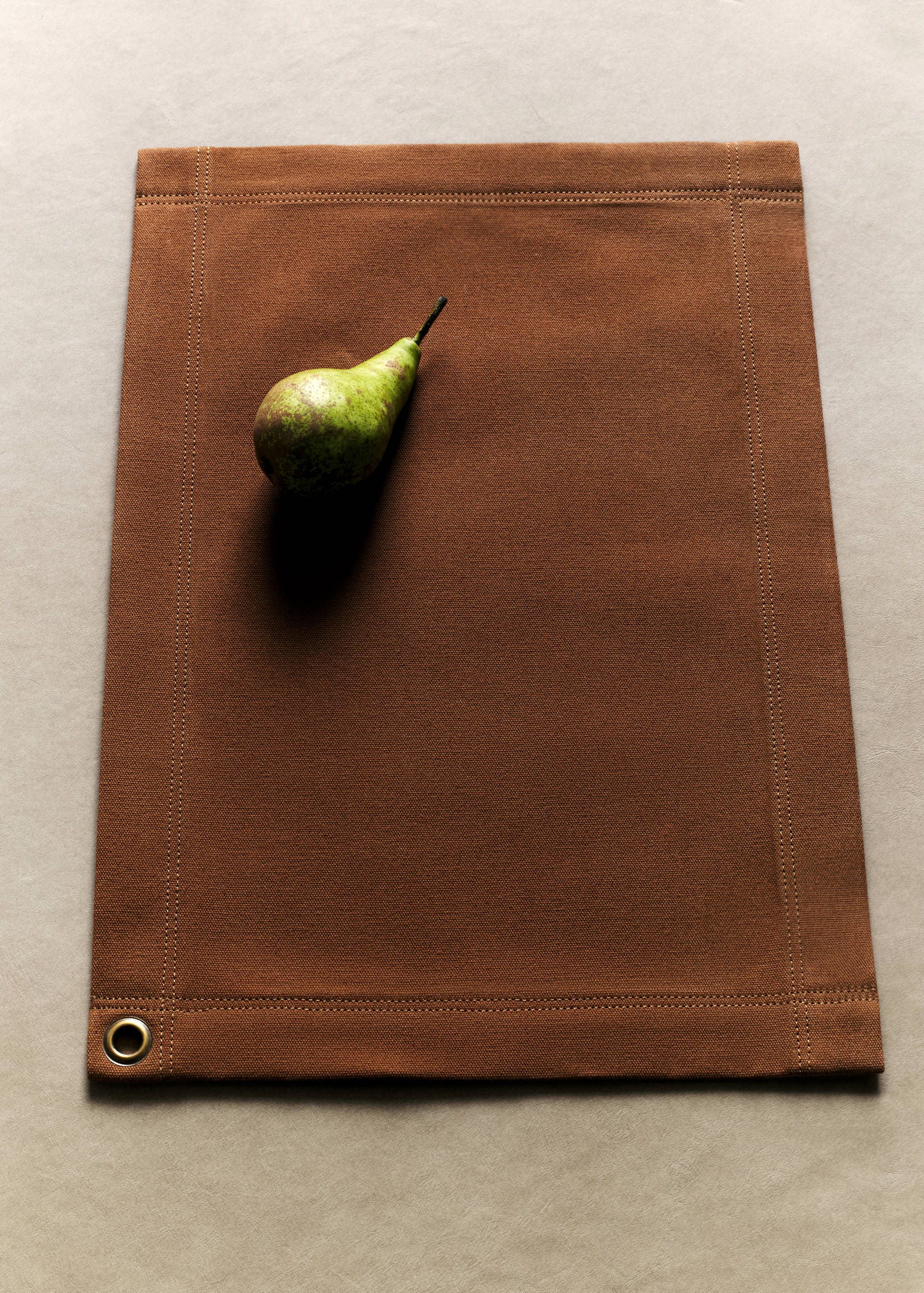 Brown placemat with topstitching and a corner eyelet, styled with a pear.
