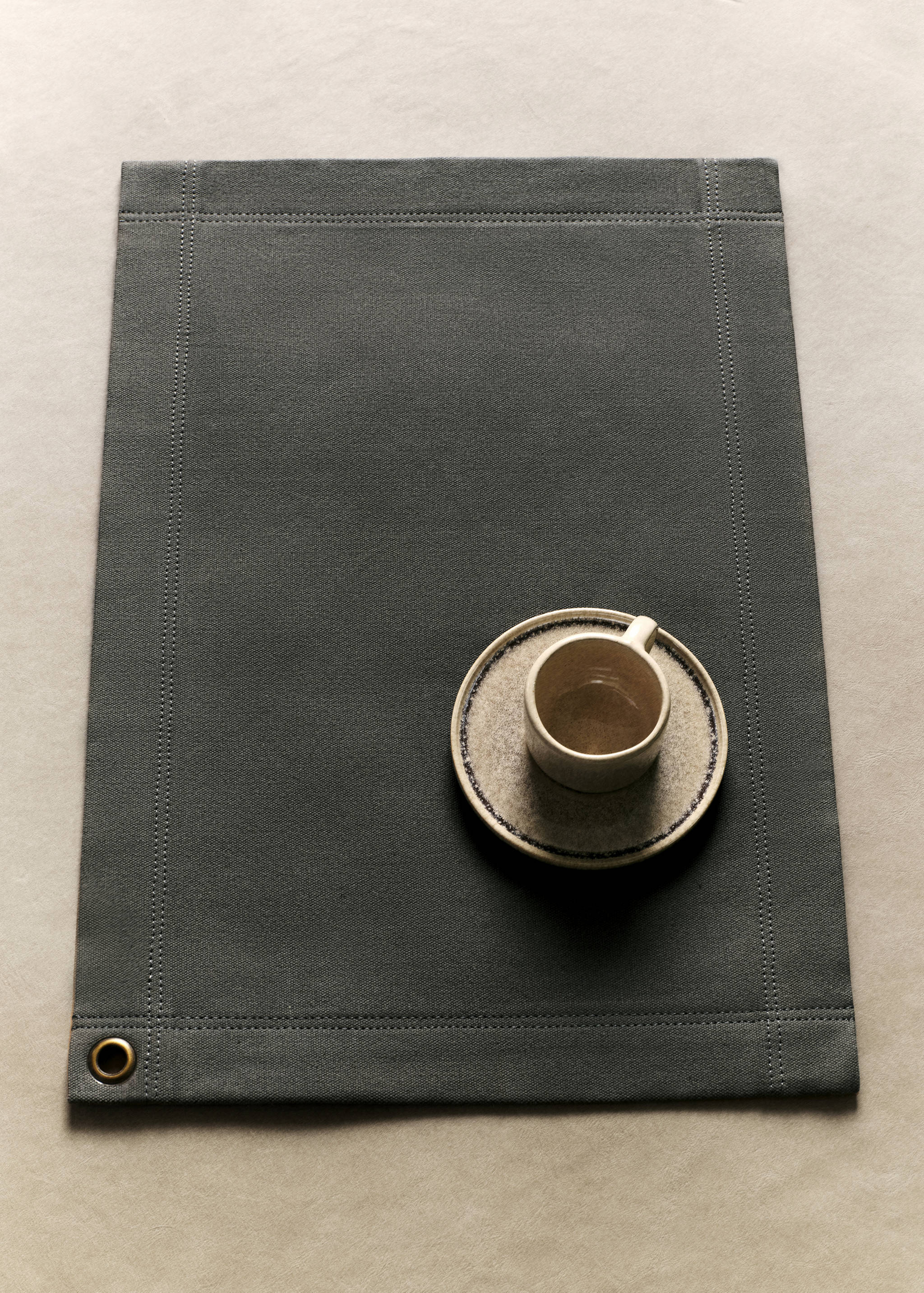 Dark grey placemat with a stitched border and brass eyelet.