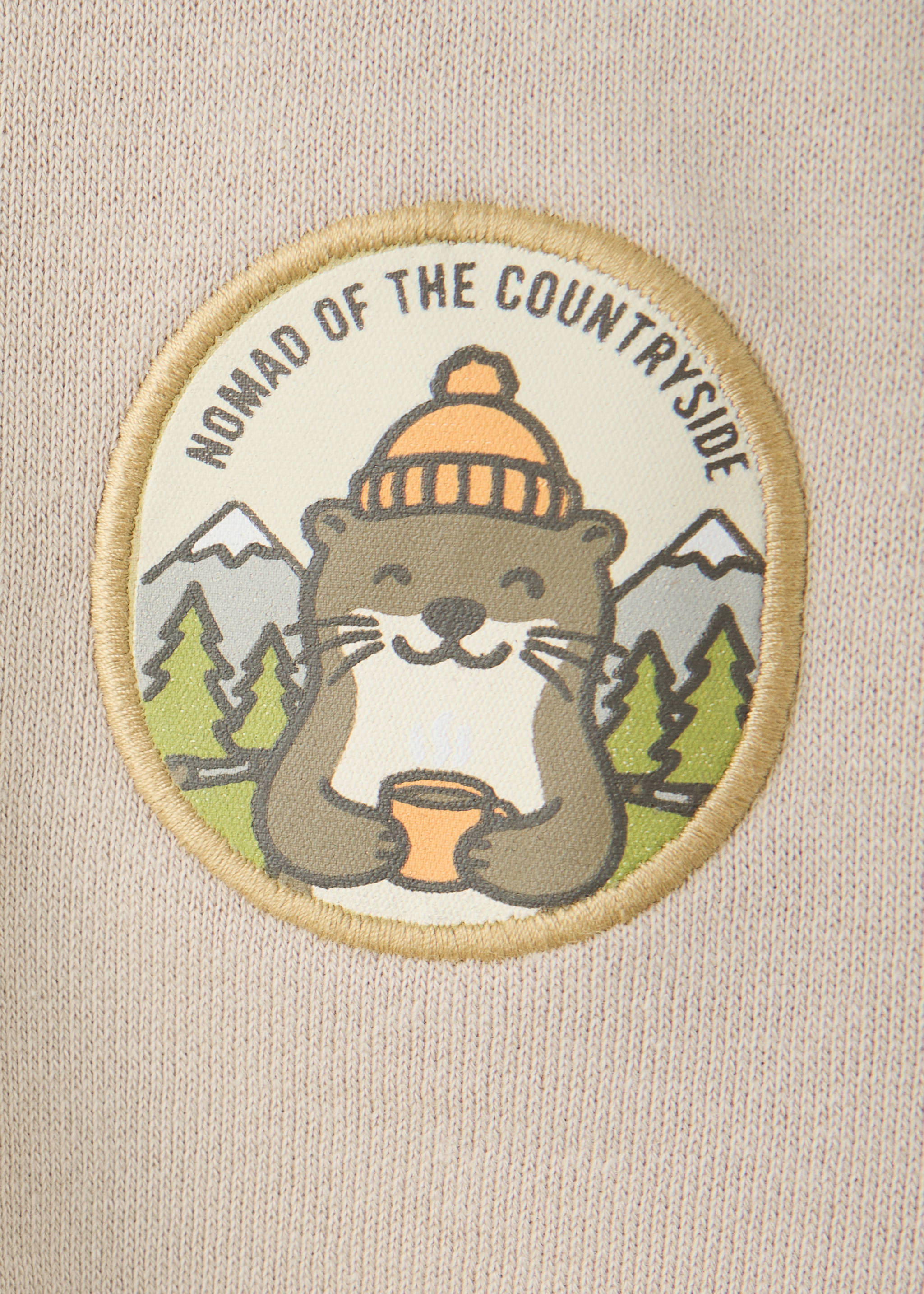 Beige sweatshirt fabric detail featuring a round otter patch with 'NOMAD OF THE COUNTRYSIDE' text.