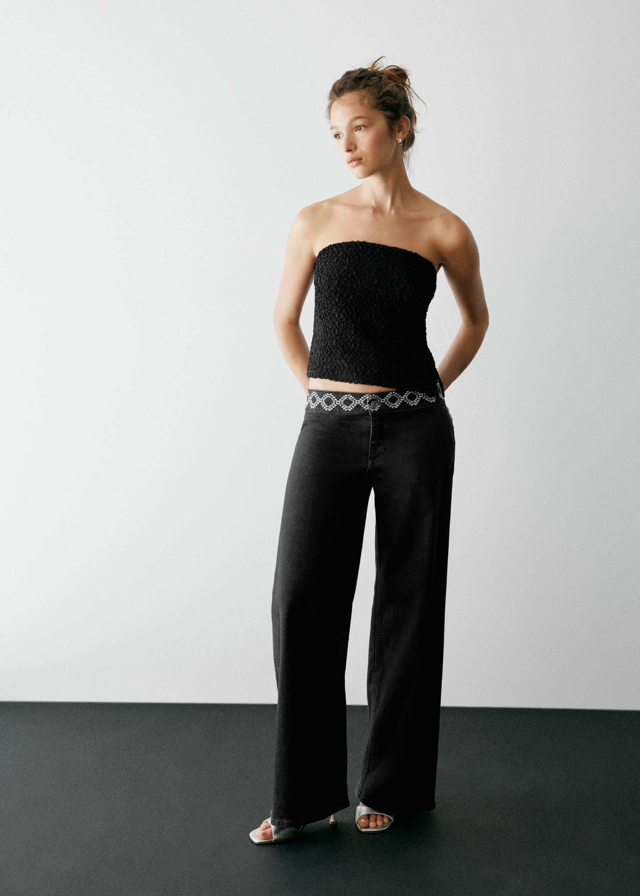 Black wide-leg denim trousers with an embellished waistband, styled with a black textured strapless top.