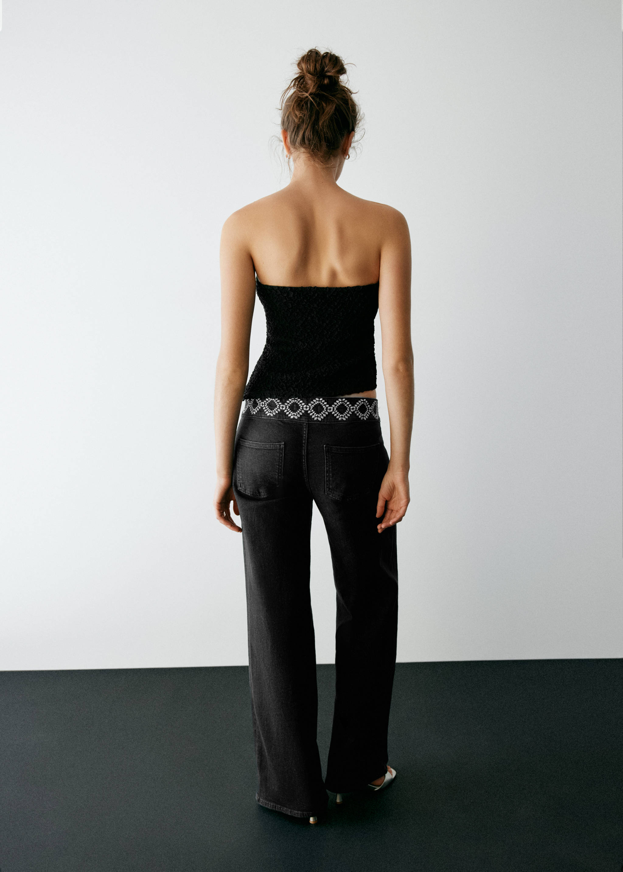 Back view of black denim trousers with an embellished waistband, worn with a black top.
