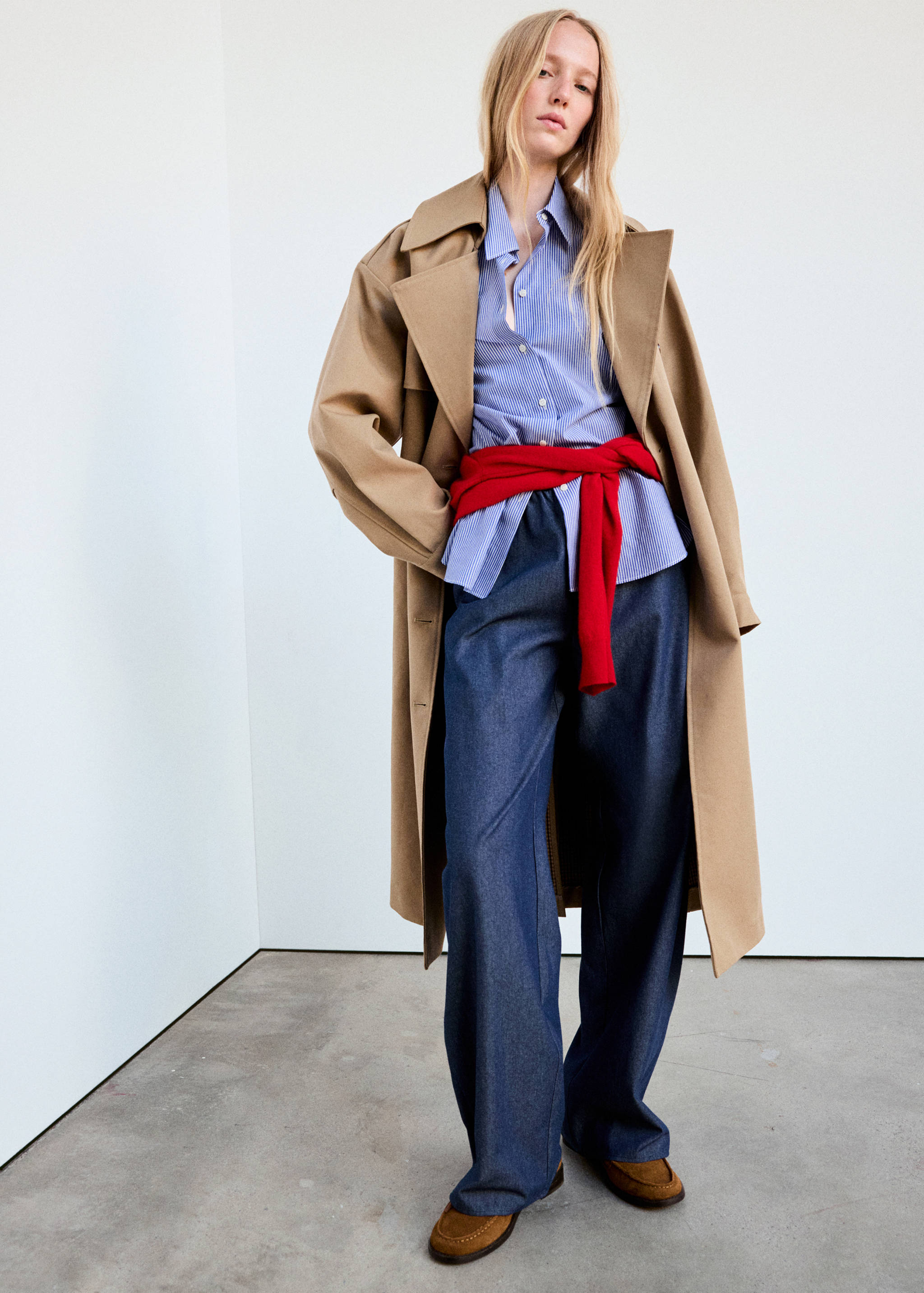 Dark blue denim trousers worn with a striped shirt, red sweater, and beige trench coat.