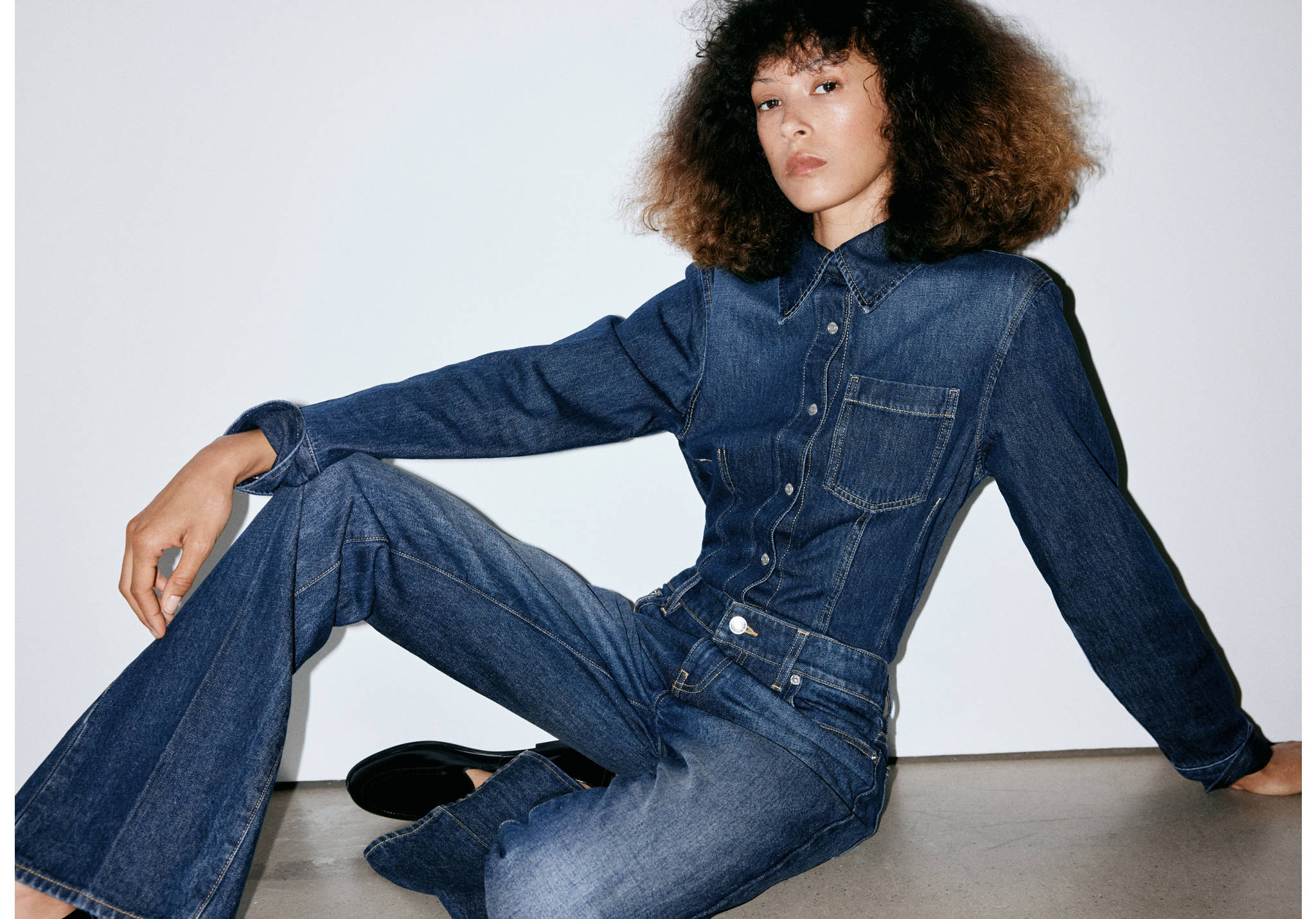 Dark blue denim jumpsuit with a pointed collar, button fastening, and flared trouser legs.