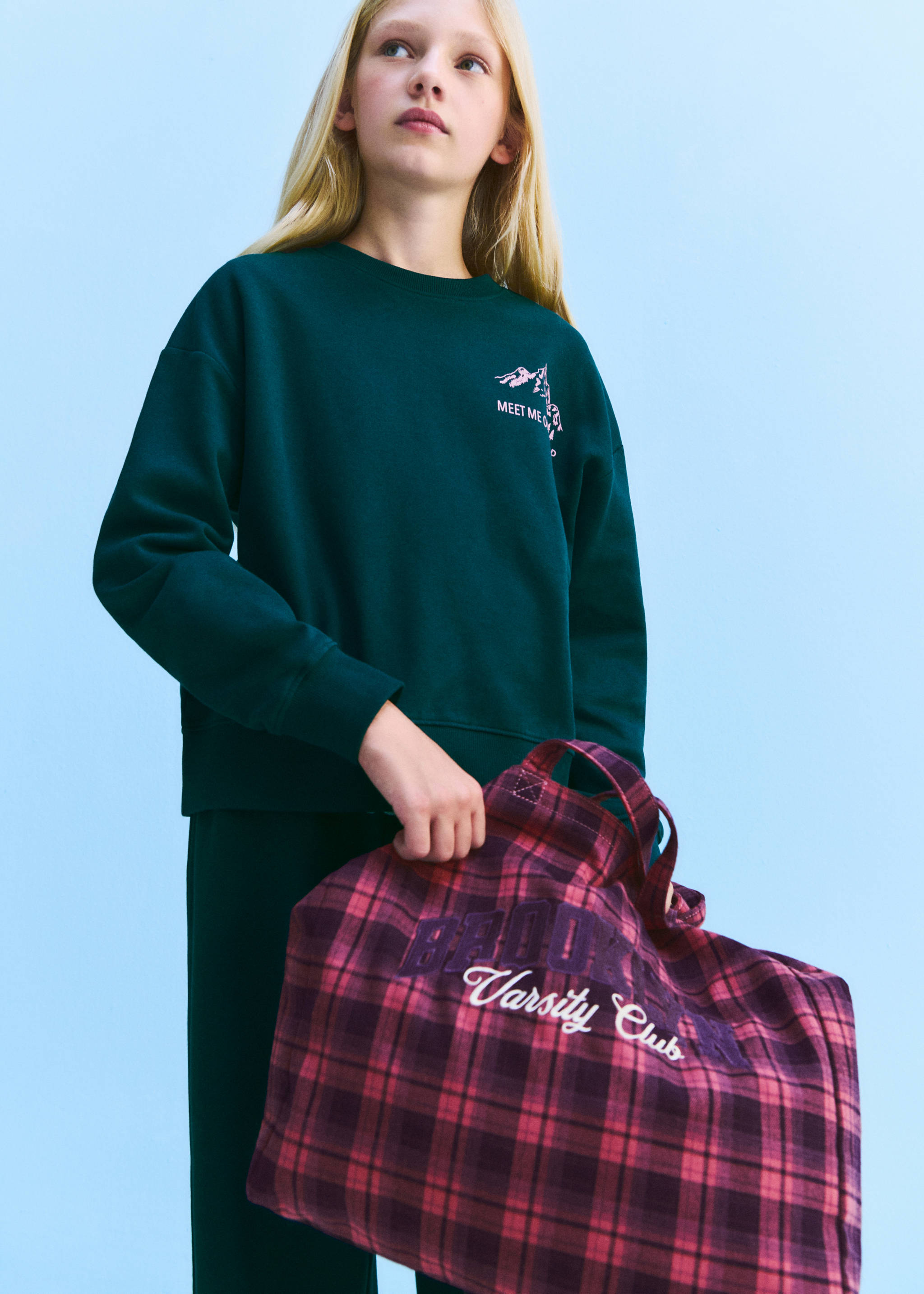 Red and dark-purple plaid tote bag with double handles and varsity lettering, held by a model.