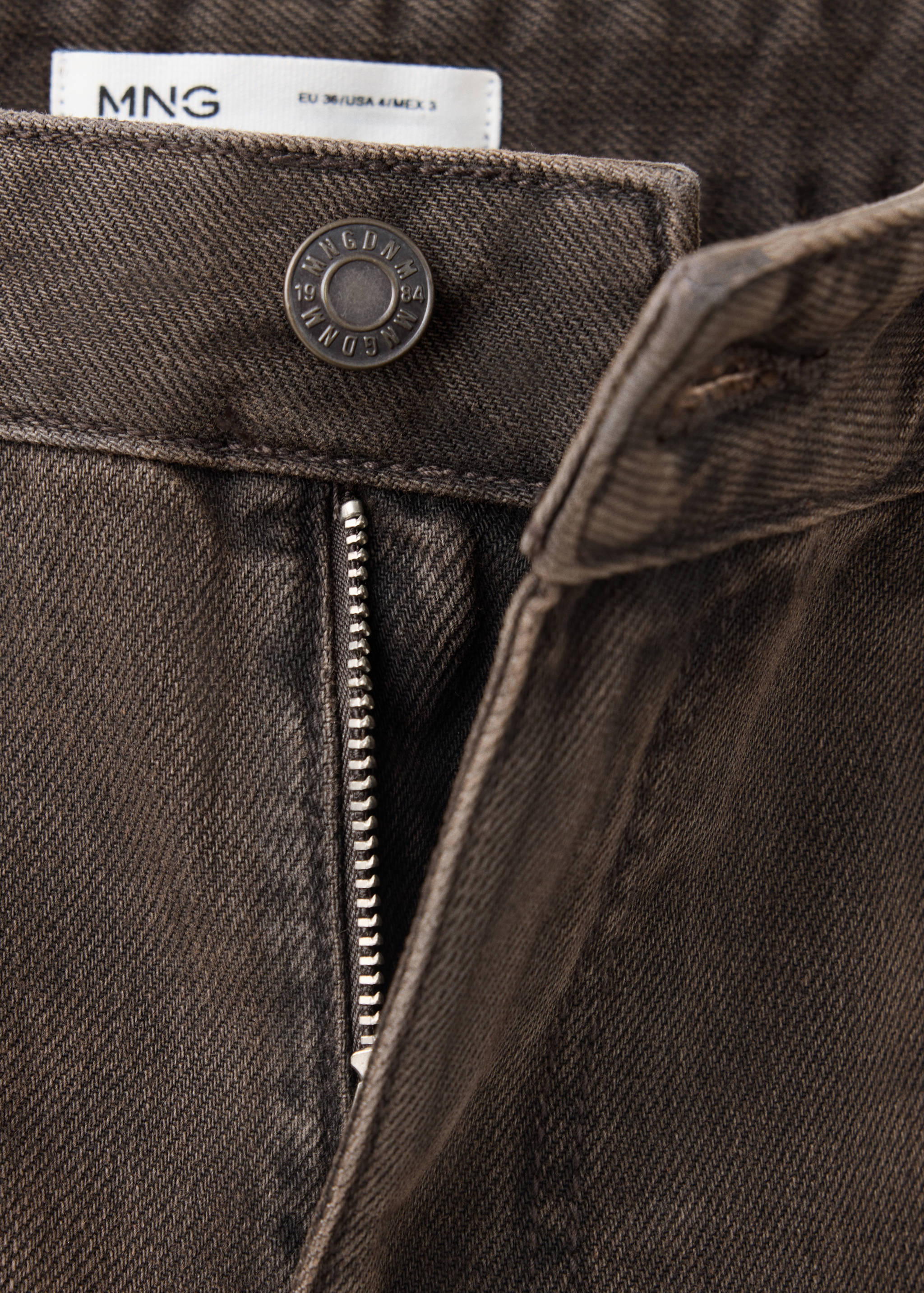 Close-up of dark brown denim trousers showing waistband, button and zip fly.