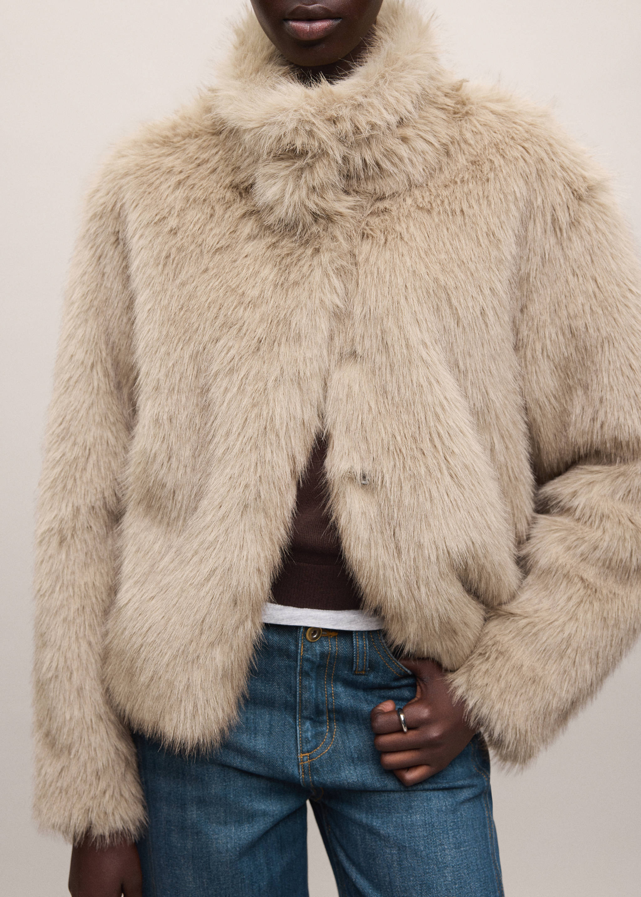 Beige faux-fur coat with a high funnel neck worn open over a brown top and blue jeans.