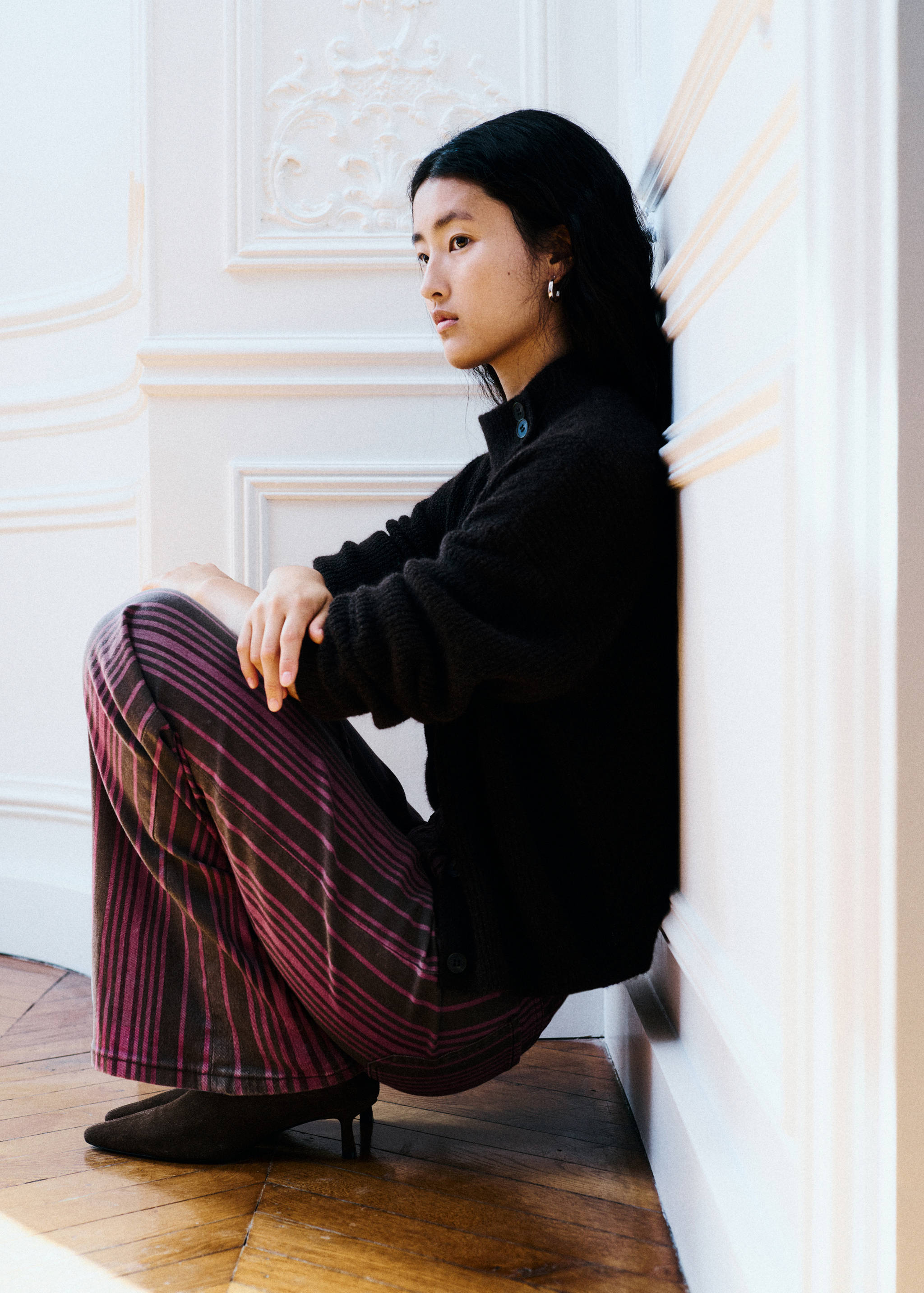 Modelo adolescente con jersey negro de punto de cuello con botones y pantalones de rayas, de perfil.