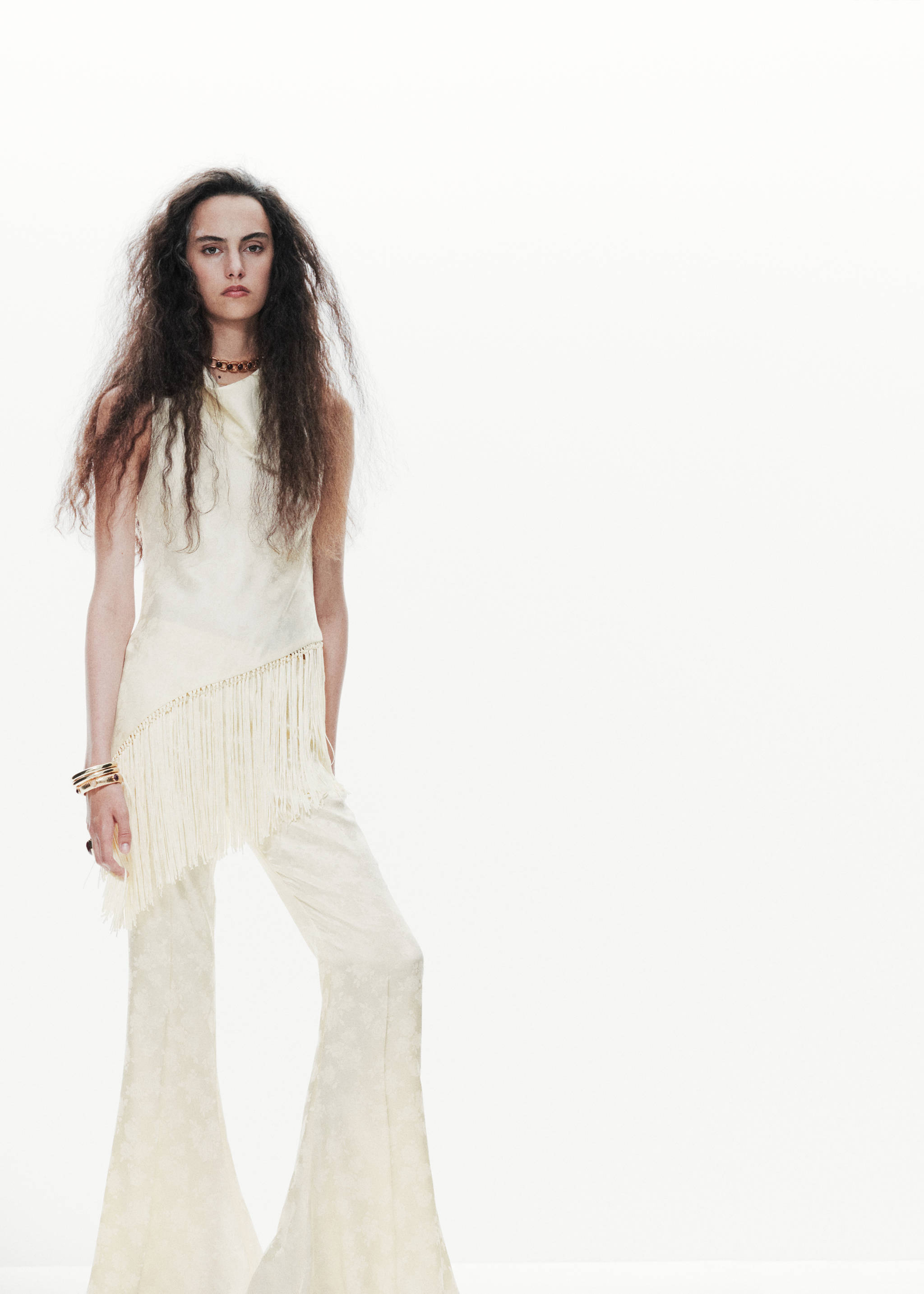 Off-white sleeveless top with an asymmetric fringe hem, worn with matching flared trousers.
