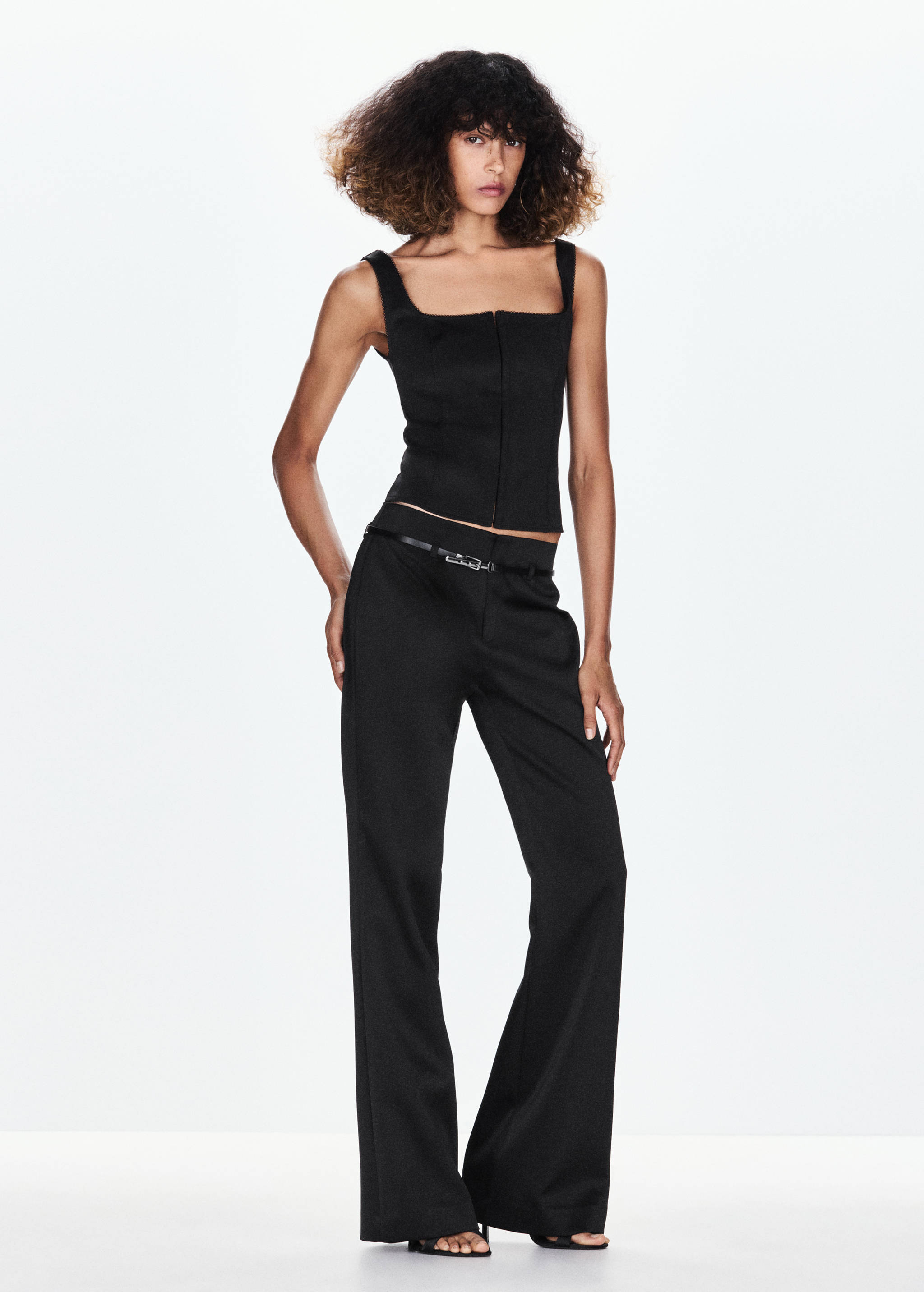 Black sleeveless waistcoat with square neckline and front zip, worn with matching trousers.