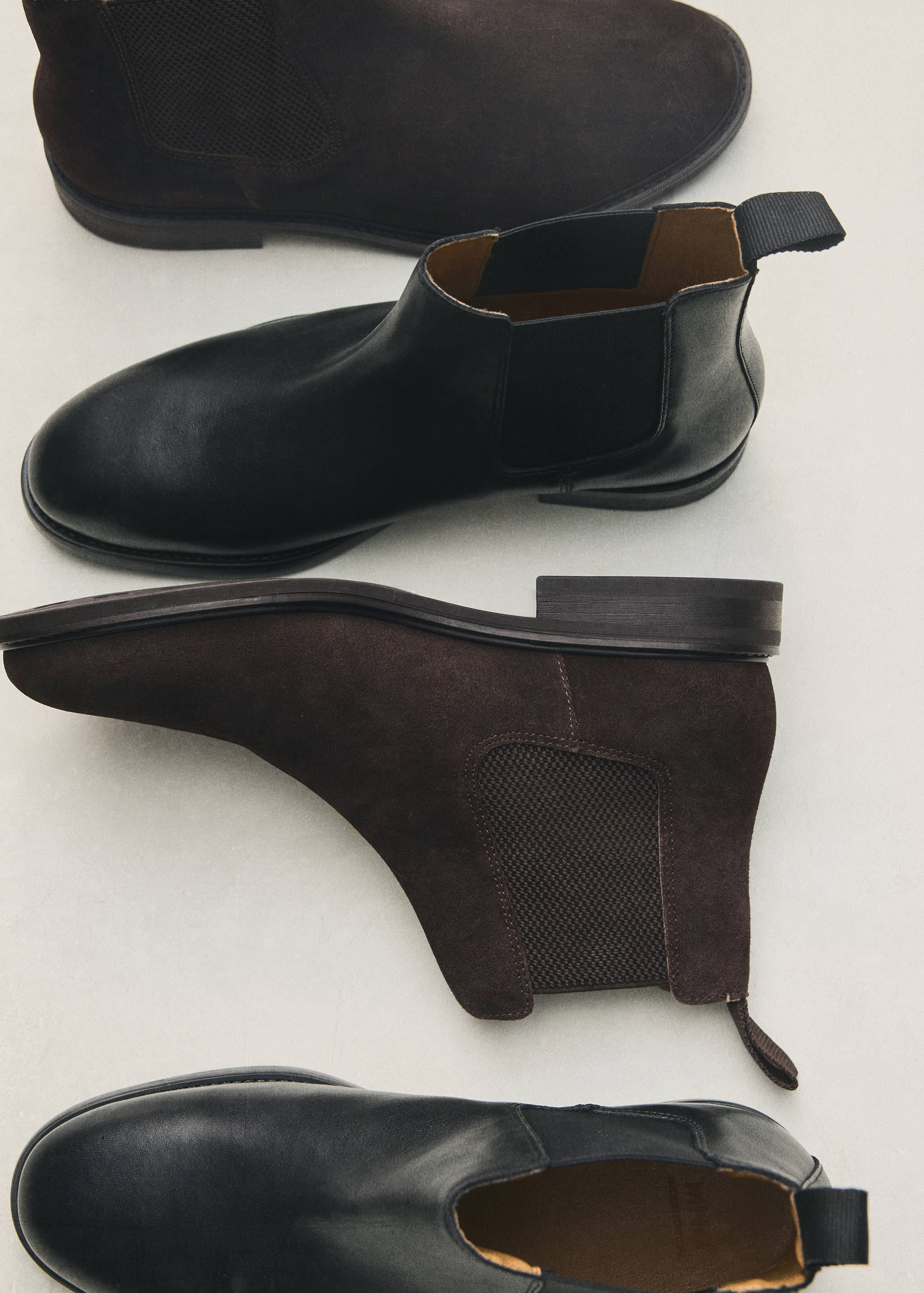 Four men's Chelsea boots in black leather and dark brown suede arranged on a light surface.