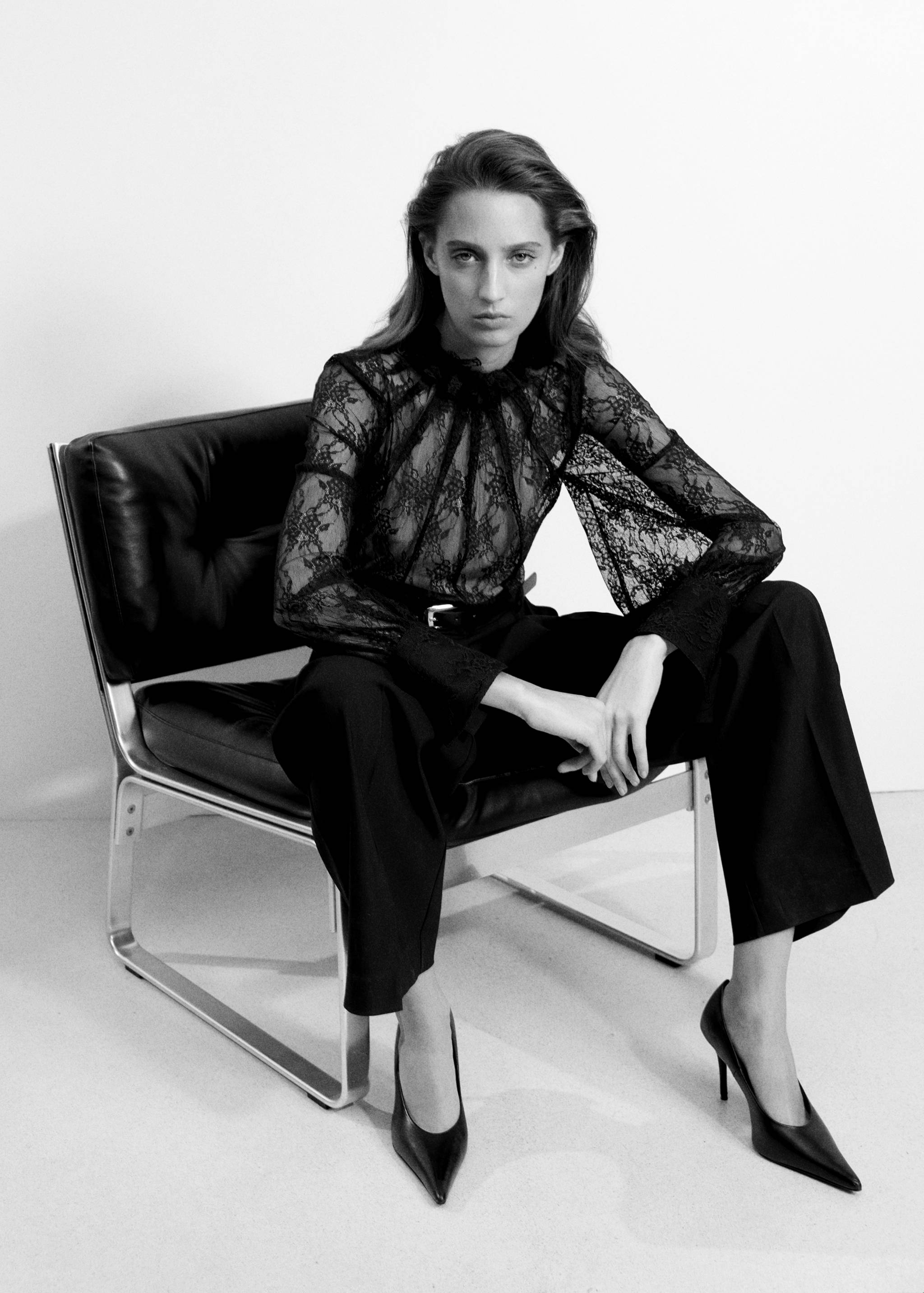 Mono largo de encaje negro con cuello alto con volantes y pantalón ancho, posando sentada.