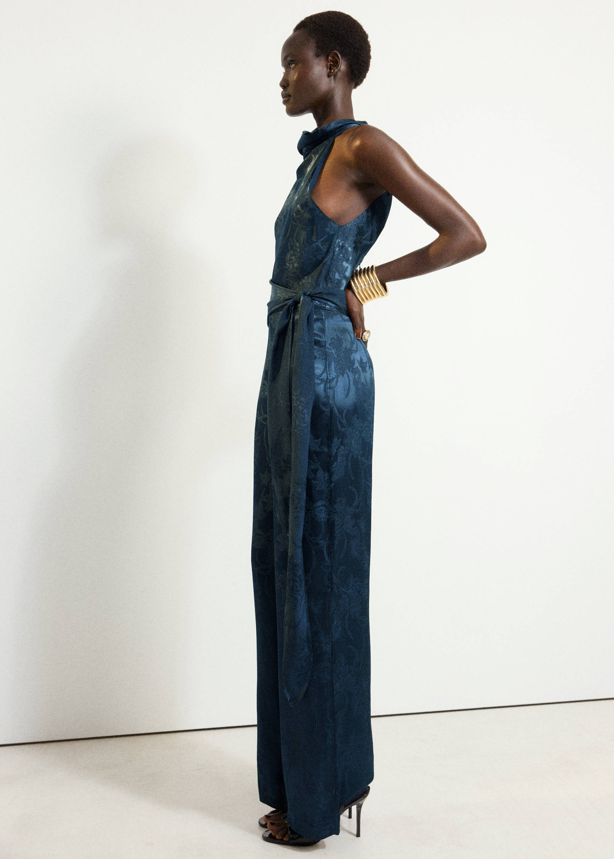 Dark teal blue satin halter neck jumpsuit with a tie belt and tonal floral pattern.