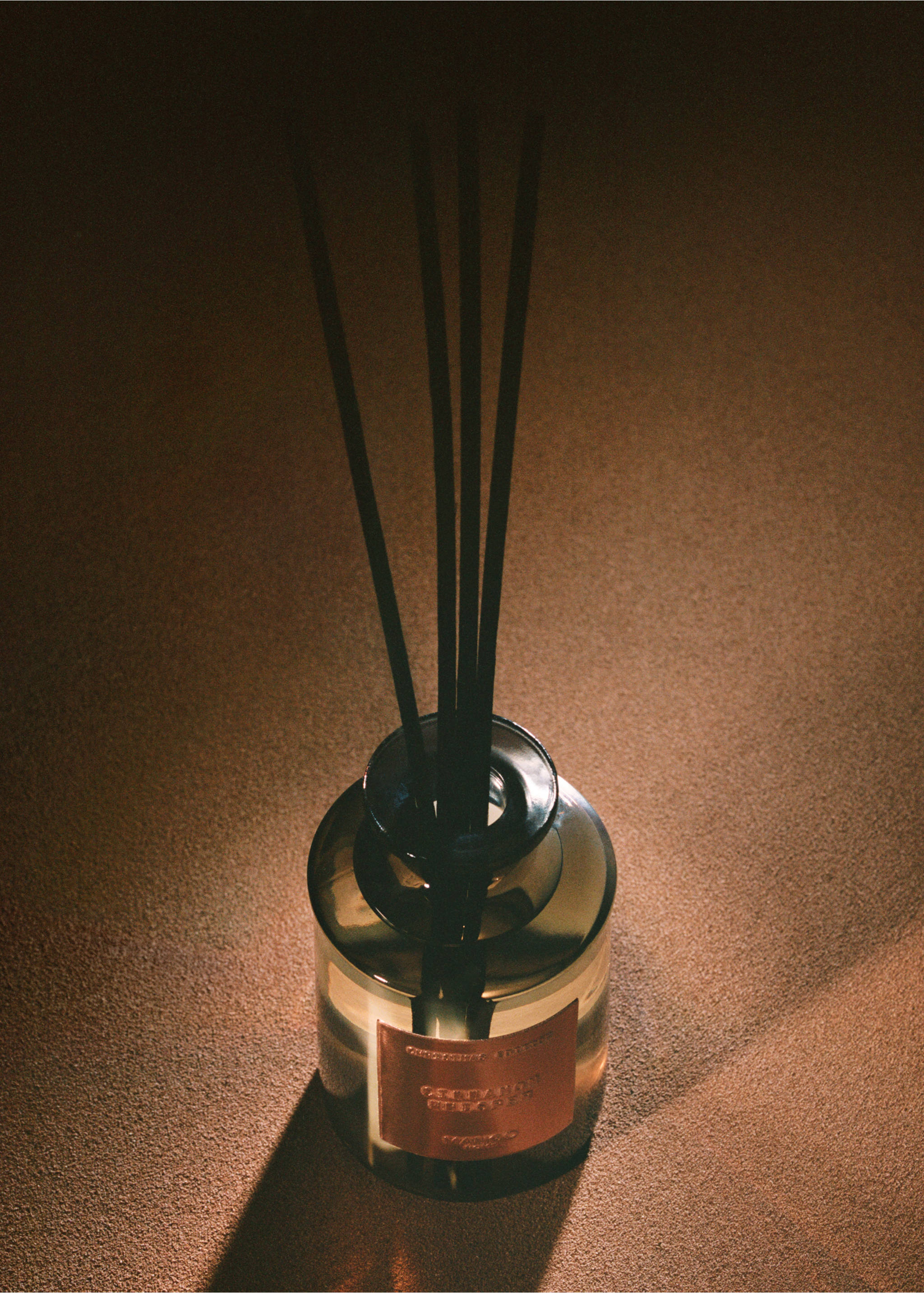 Smoked-glass reed diffuser with black sticks on a textured brown surface., Copper. Ref: 37087810-00.