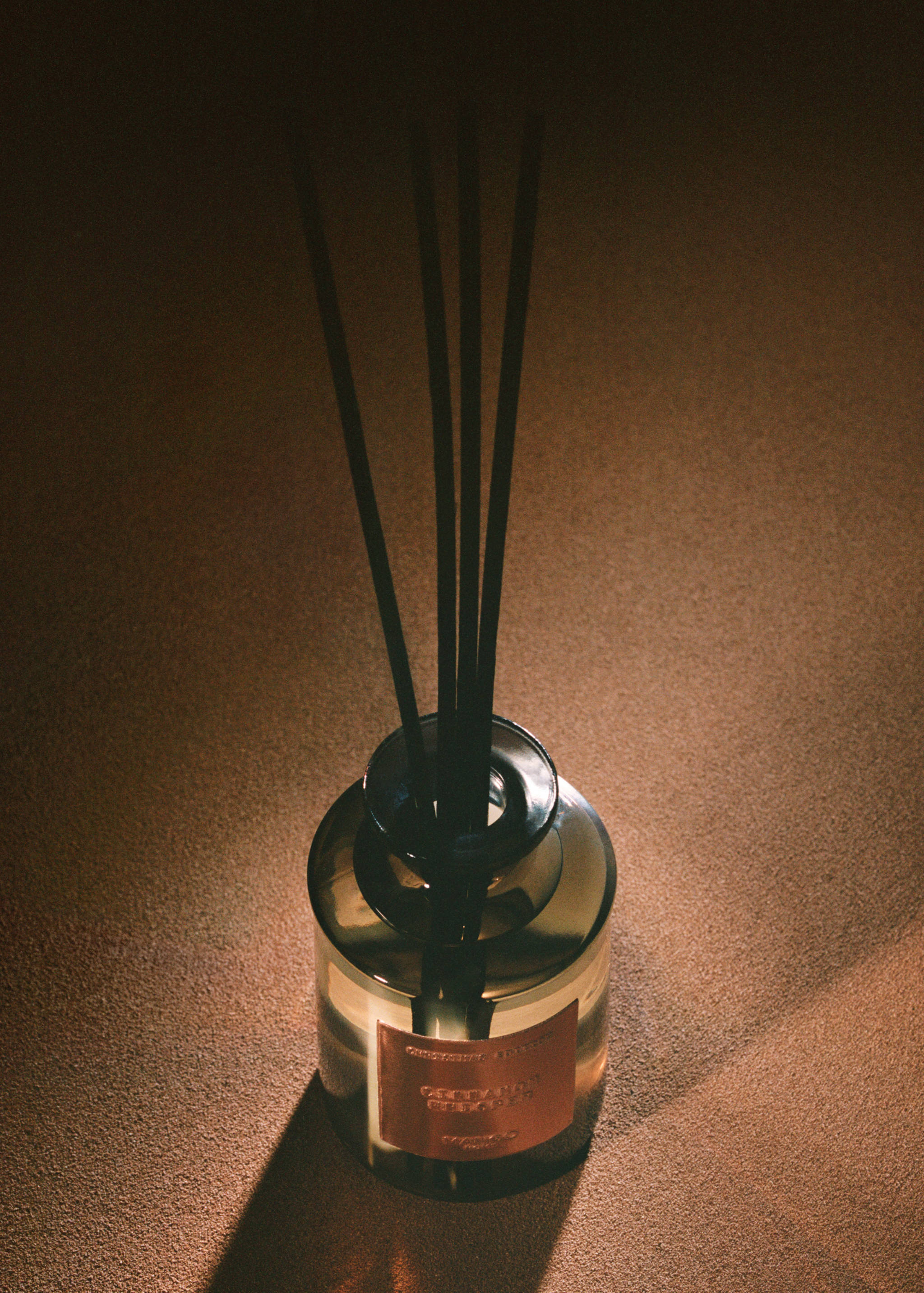 Smoked-glass reed diffuser with black sticks on a textured brown surface.