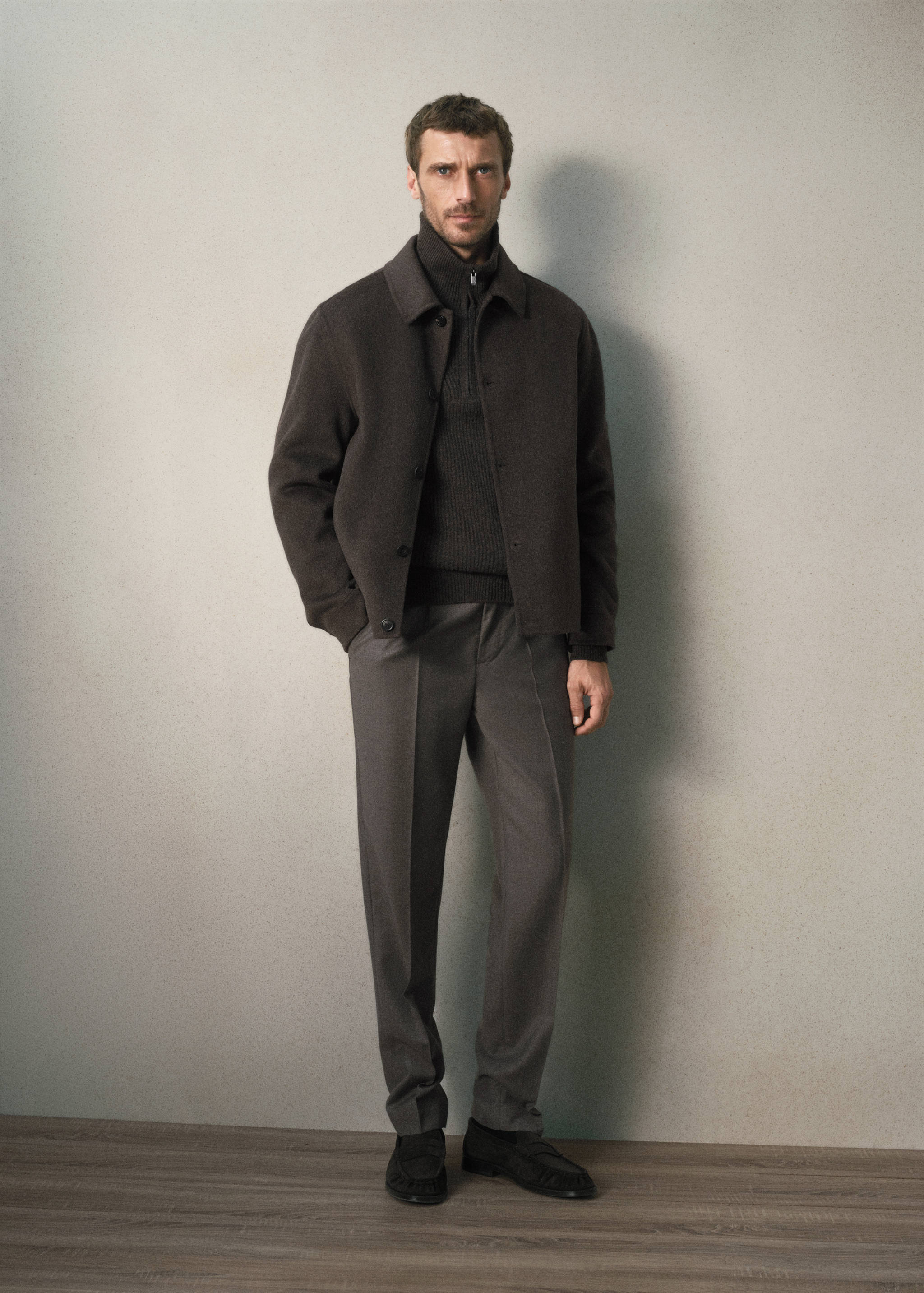 Dark brown jacket with classic collar and button fastening, worn with a dark grey jumper and trousers.
