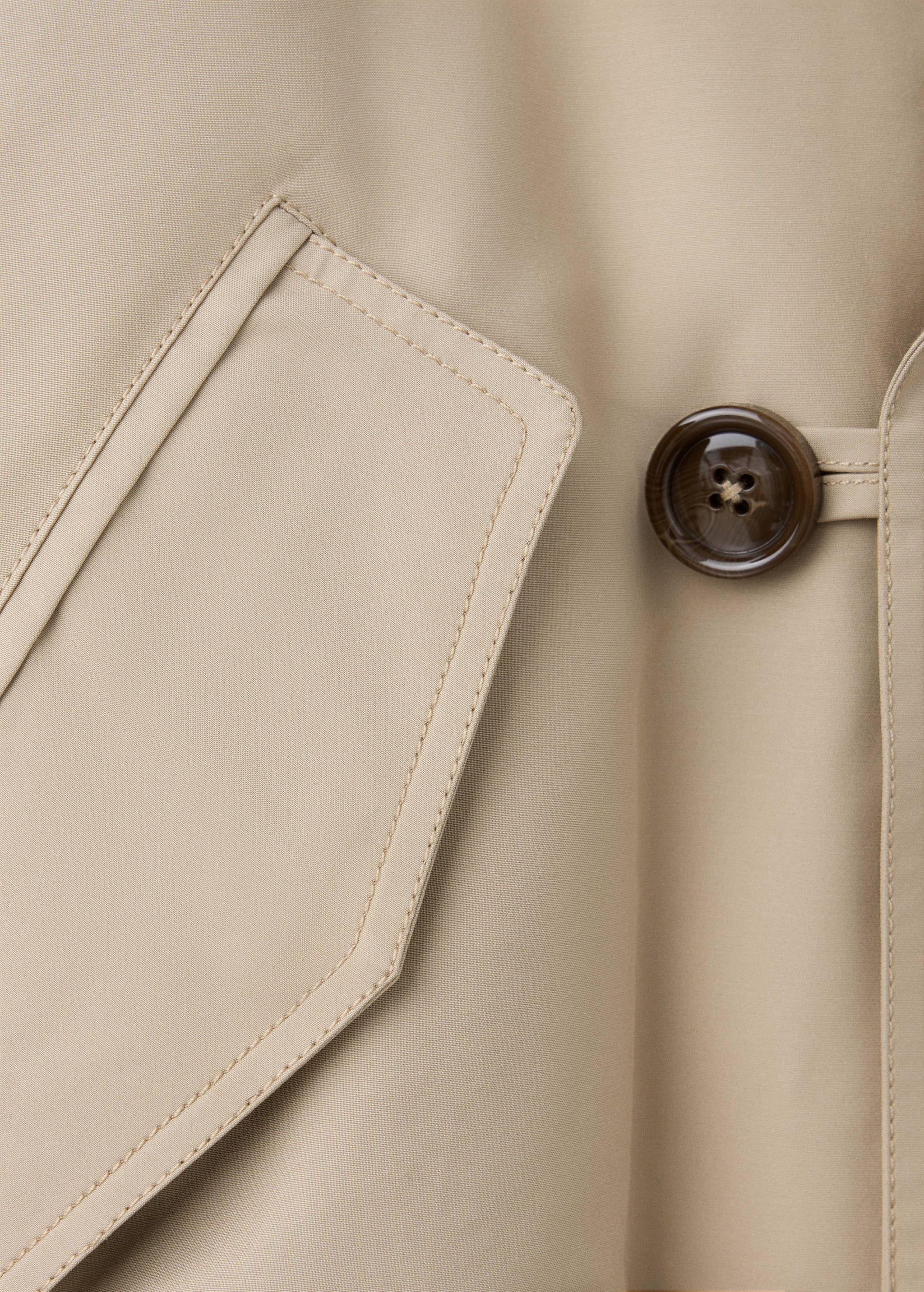 Close-up of a beige jacket showing the fabric detail, pocket flap, and tortoiseshell button.