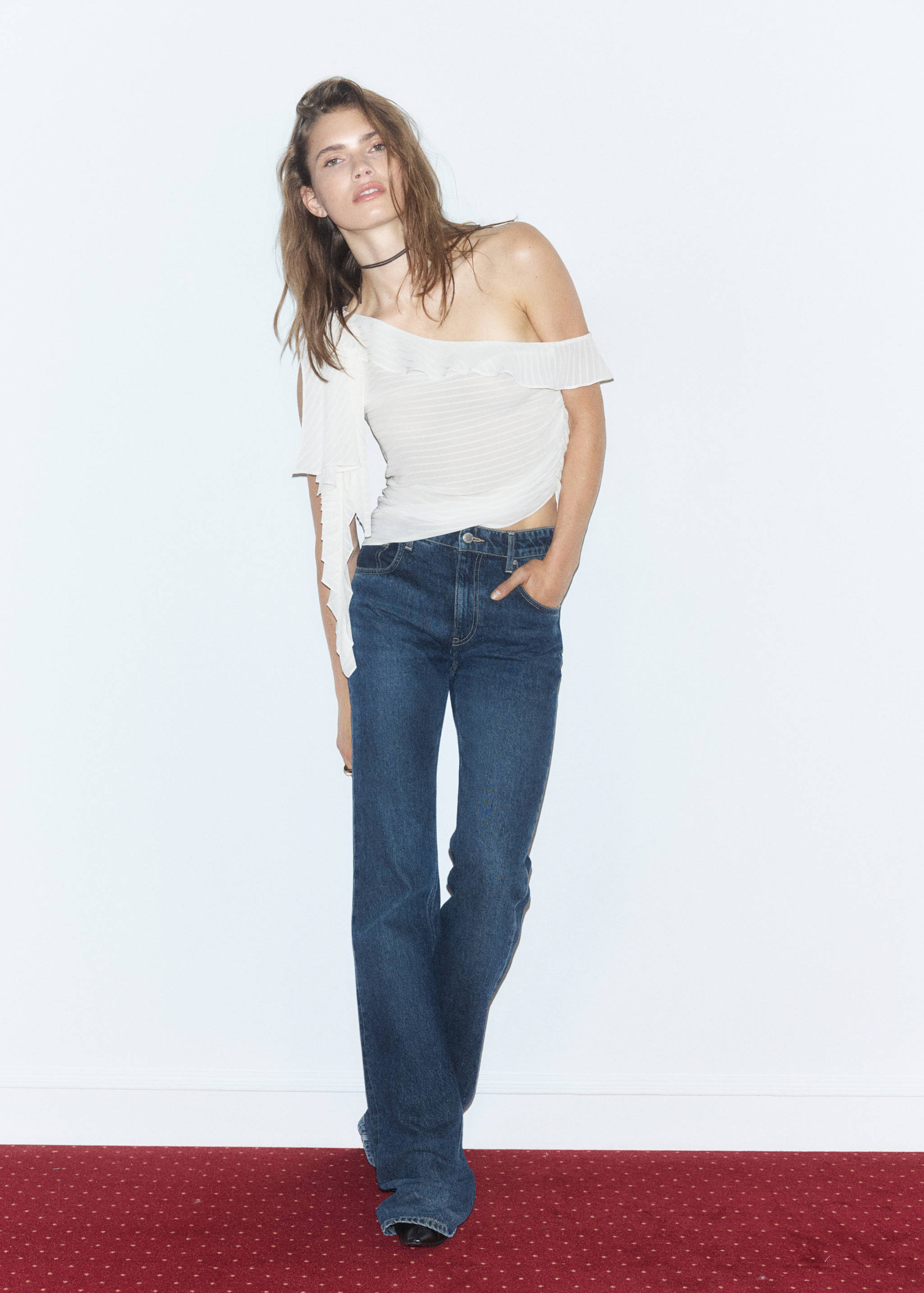 White off-the-shoulder pleated top with ruffles, worn with blue denim trousers.