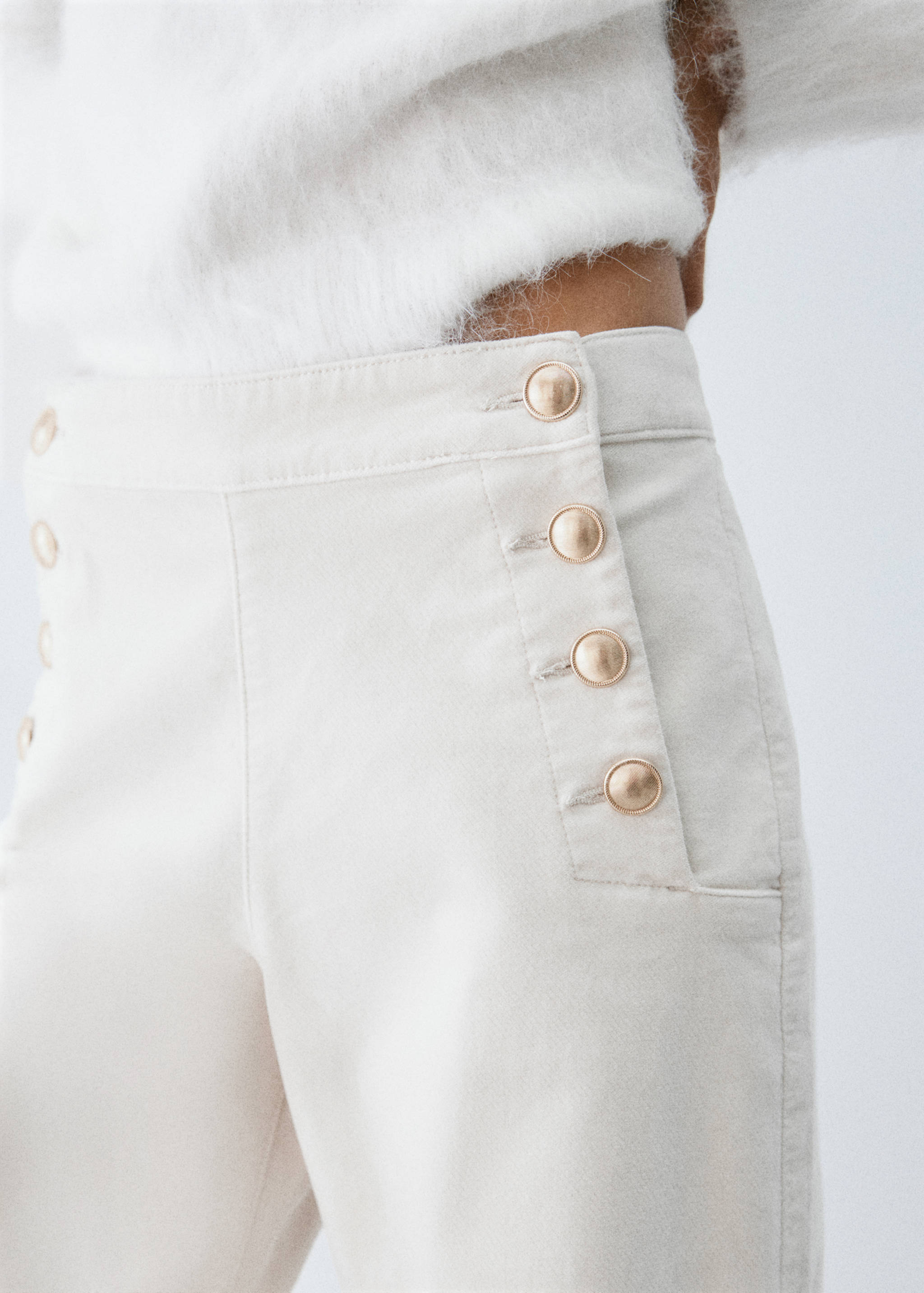 Off-white denim trousers with gold buttons, paired with a white knit sweater.