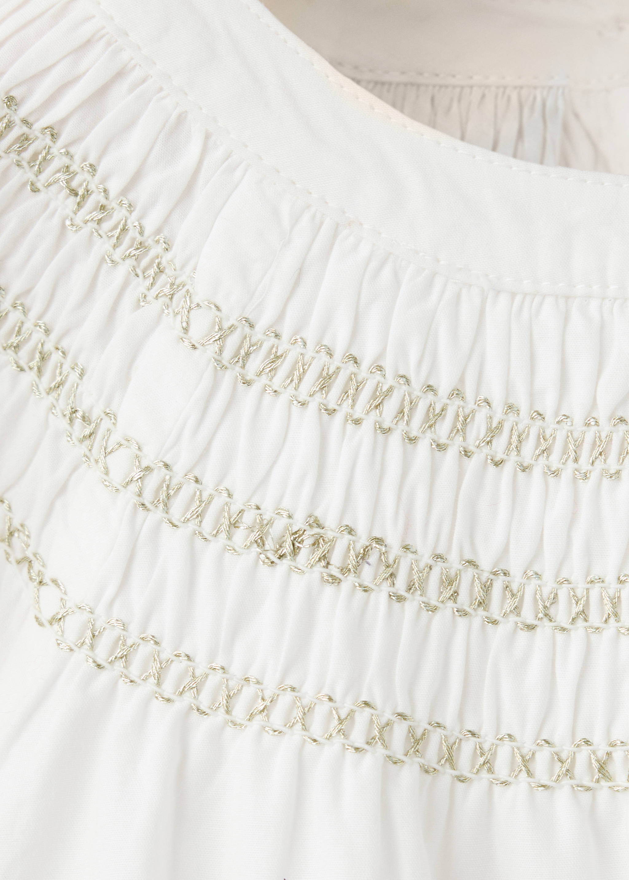 Detalle de blusa infantil blanca con nido de abeja dorado.
