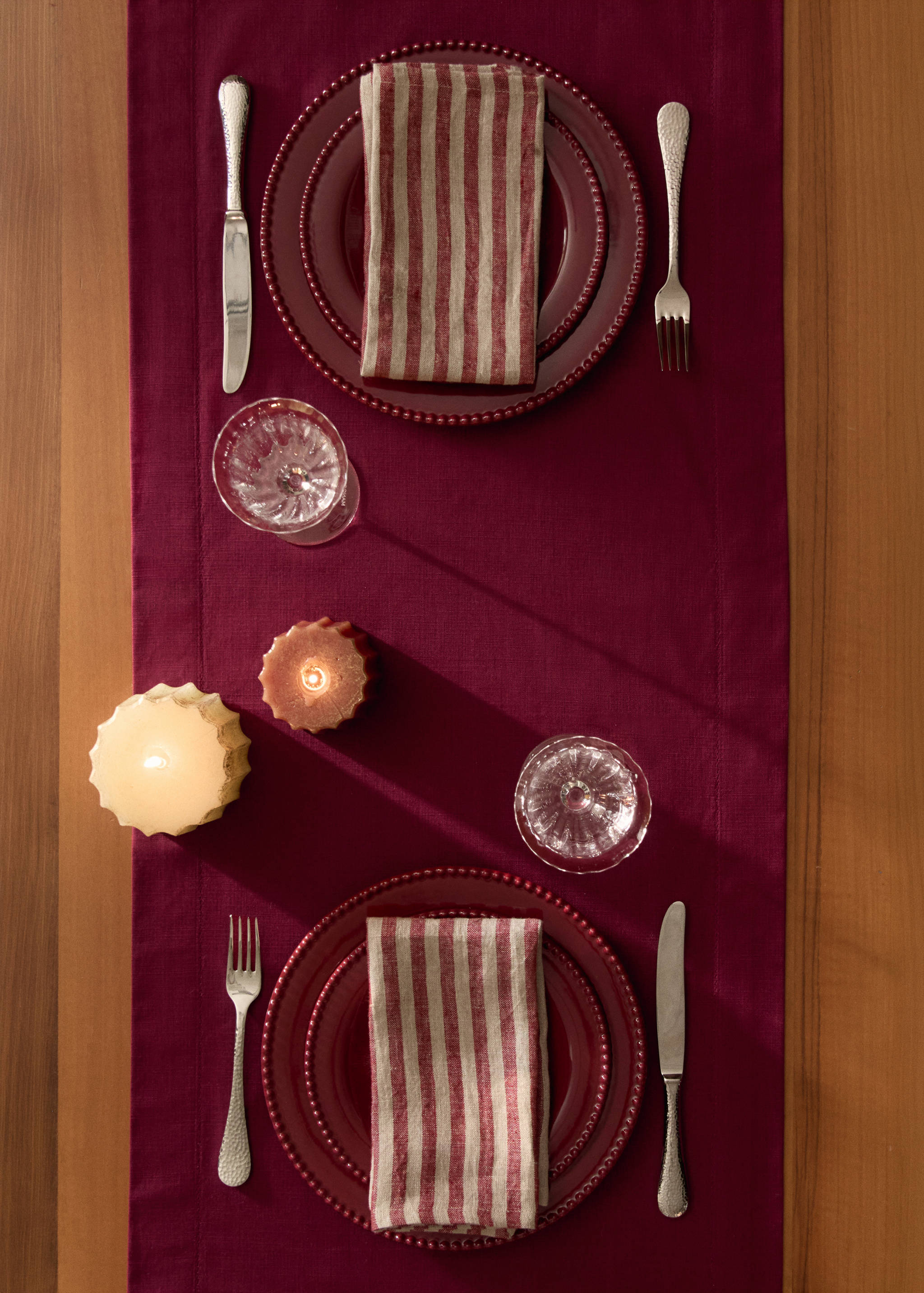 Burgundy table runner in an overhead table setting with plates, striped napkins, silverware, and lit candles.