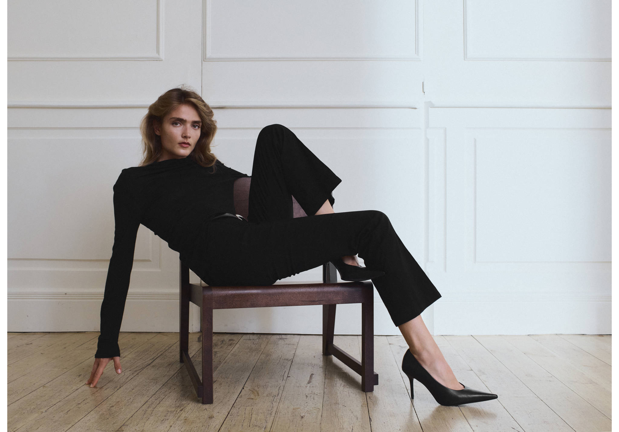 Pantalones negros puestos por una modelo sentada frente a una pared de paneles blancos.