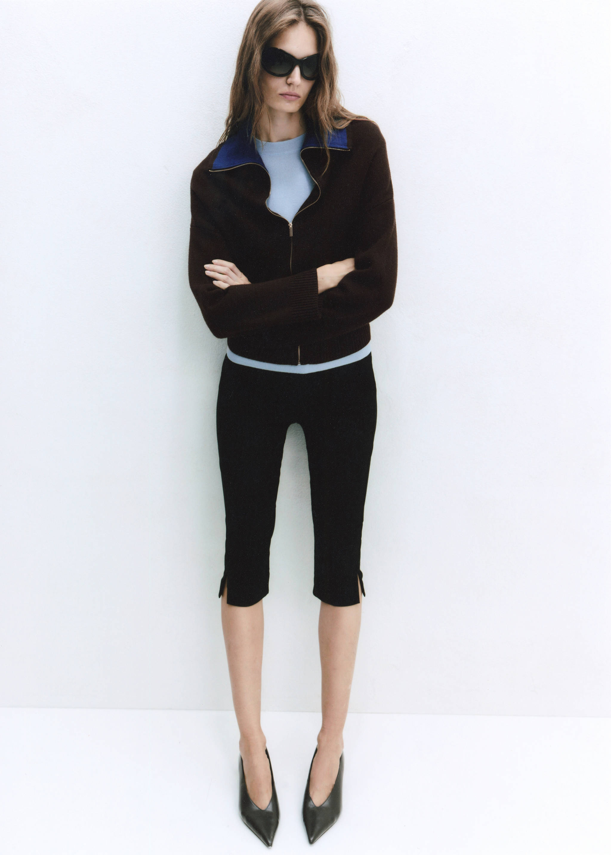 Brown zip-up jacket with contrasting blue collar, worn with a light blue top and black cropped trousers.