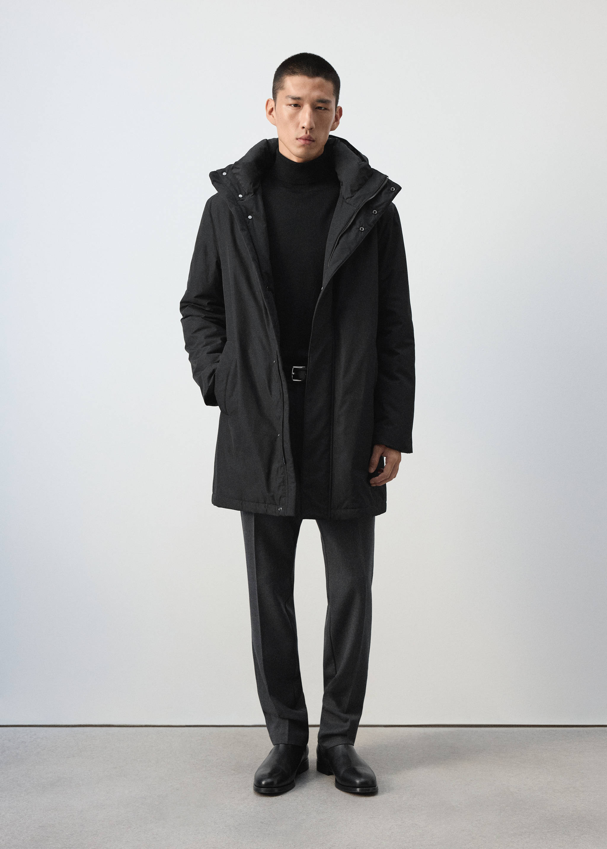 Black hooded parka worn over a black turtleneck and dark grey trousers.