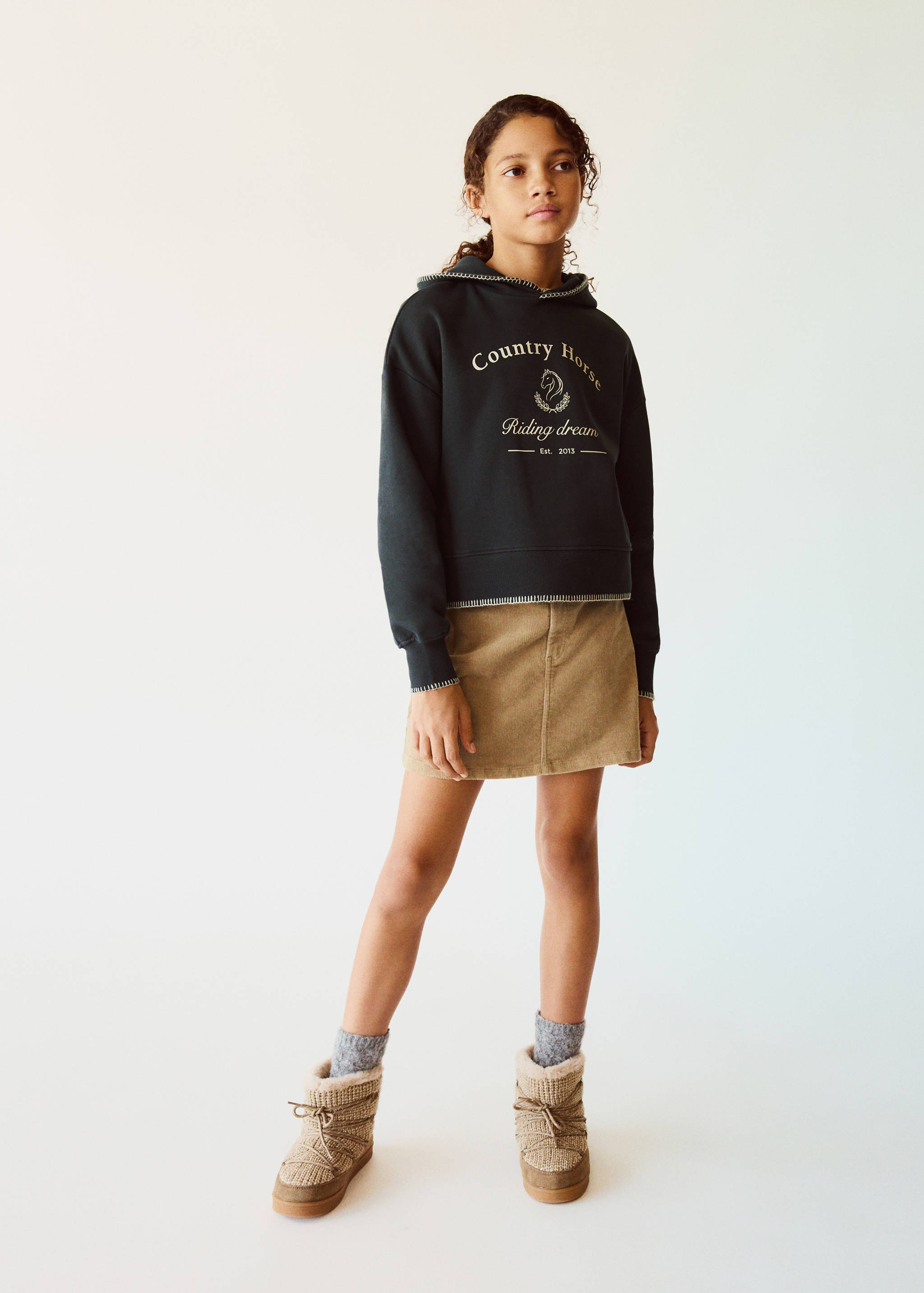 Dark green hooded sweatshirt with contrast stitching and graphic text, worn with a beige corduroy skirt.