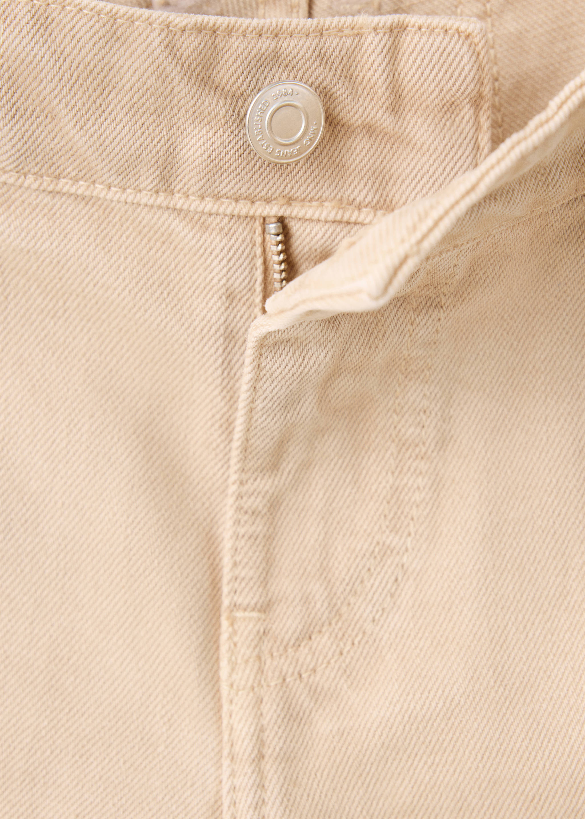Close-up of beige denim trousers showing the waistband, button, and zipper detail.
