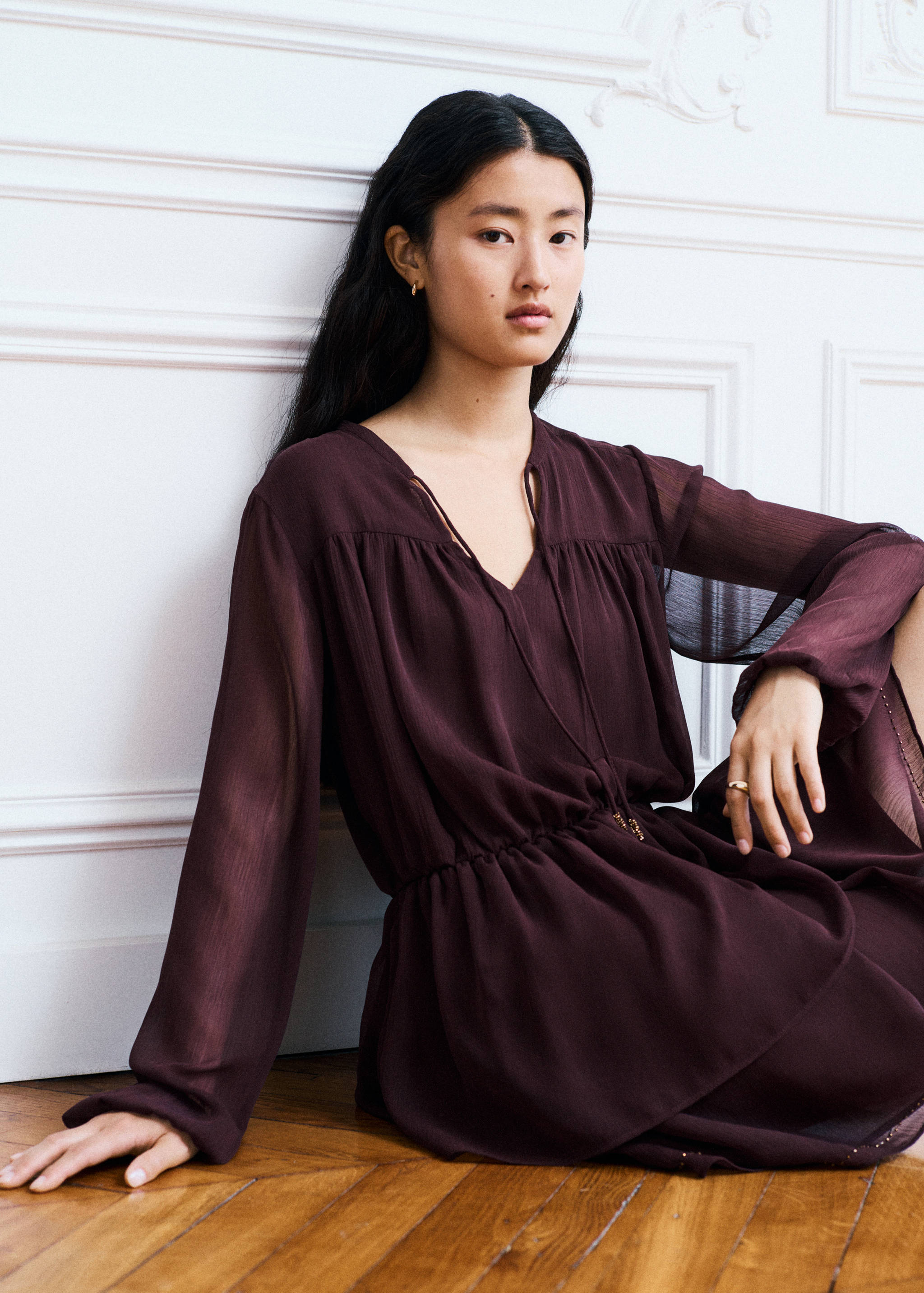 Plum sheer long-sleeved dress with a V-neck and tie closure, worn seated.
