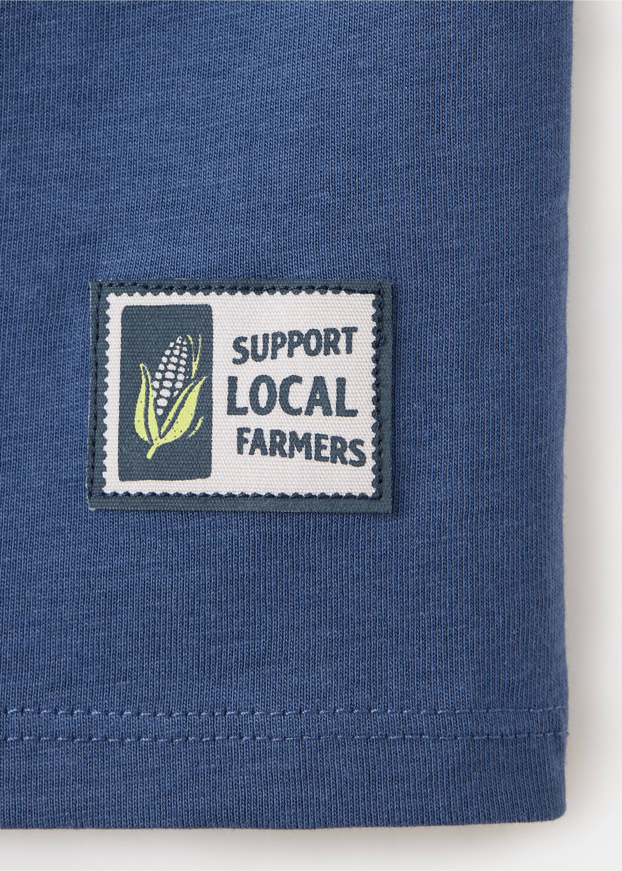 Close-up of a blue t-shirt fabric featuring a patch with corn graphic and text., Blue. Ref: 37098631-00.
