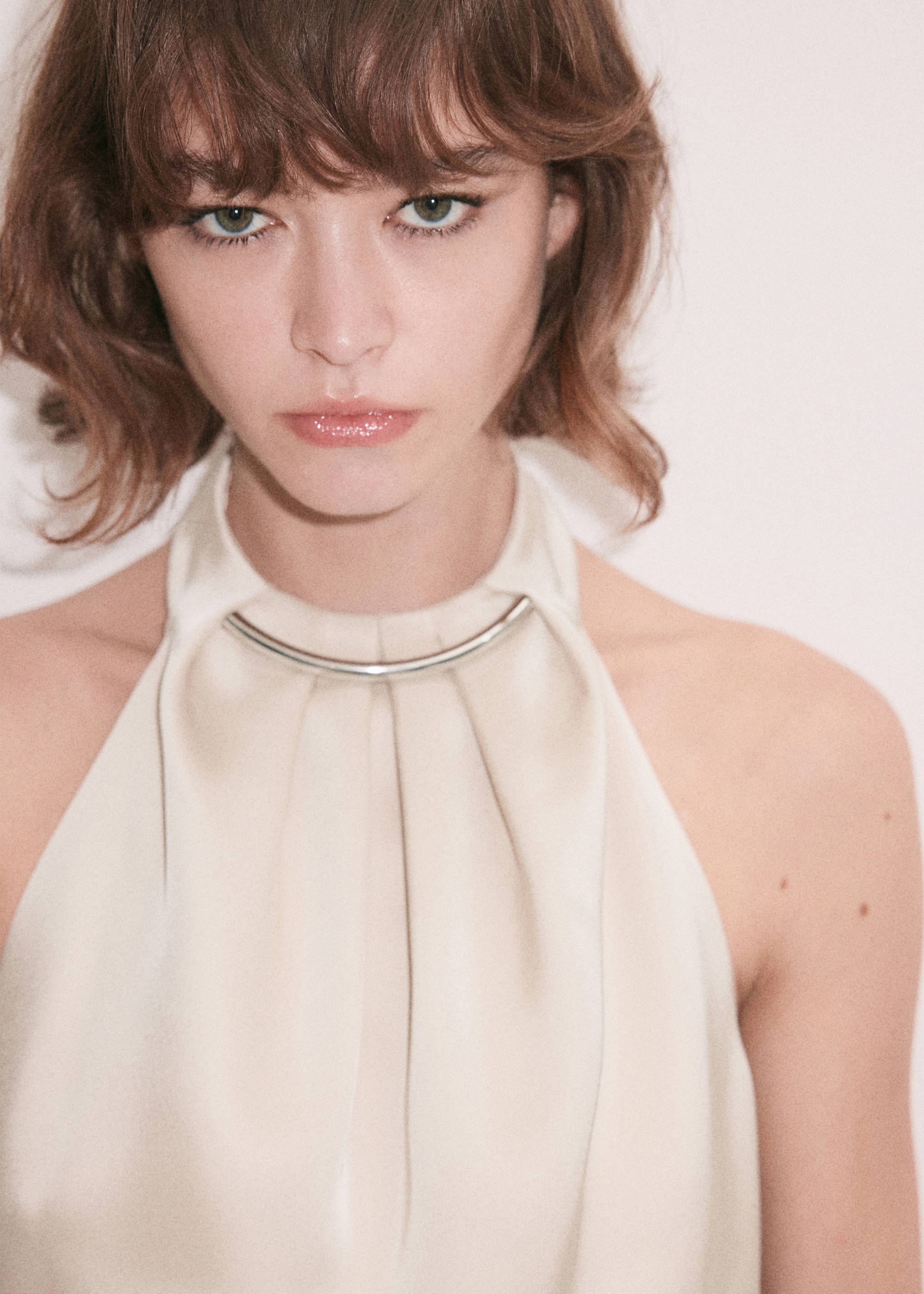 Close-up of a cream halter-neck top with pleated drape and a metallic neck bar.
