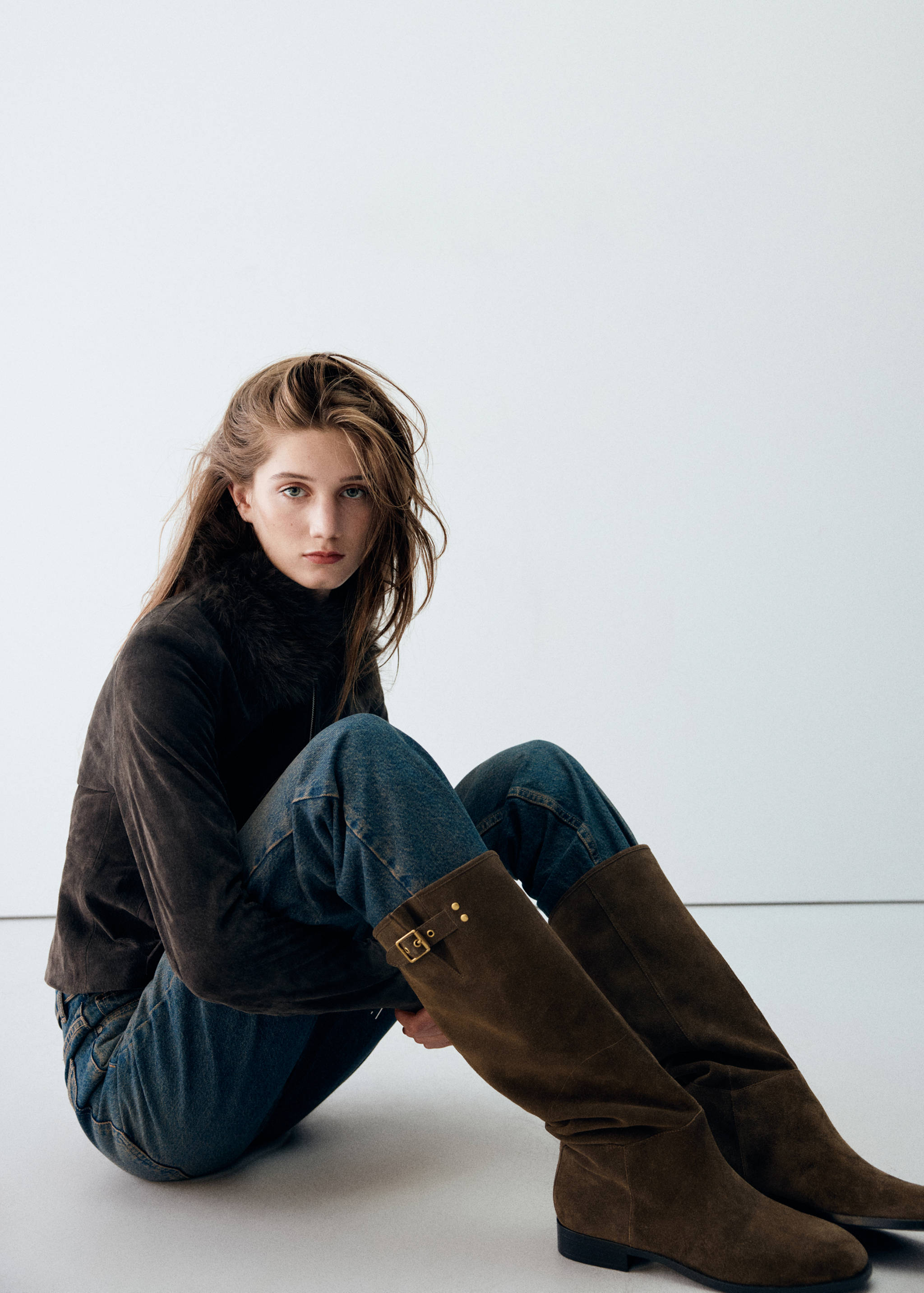 Dark brown suede knee-high boots with gold buckles, worn with blue jeans.