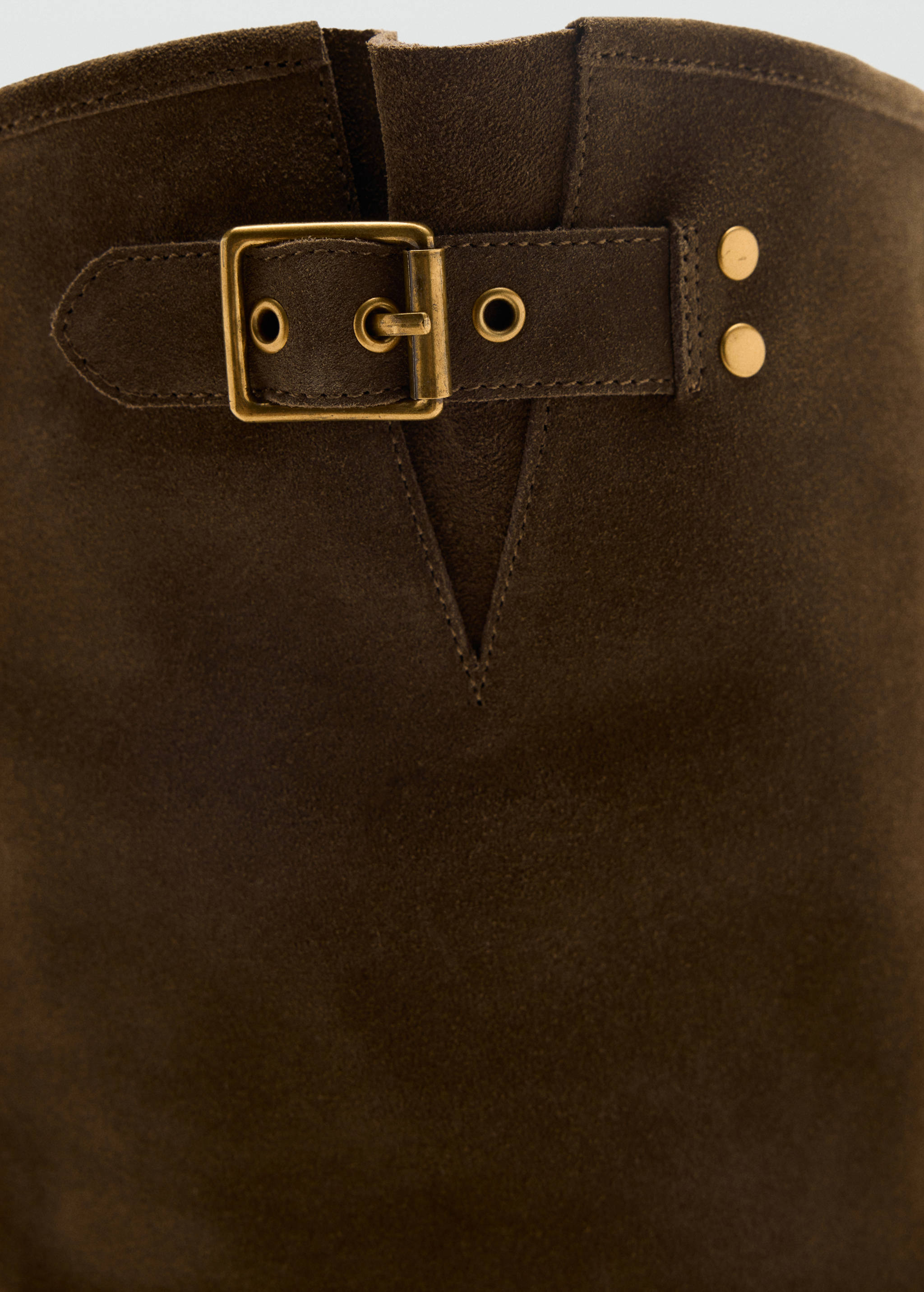 Close-up of a dark brown suede boot shaft featuring a buckled strap with antique brass hardware.