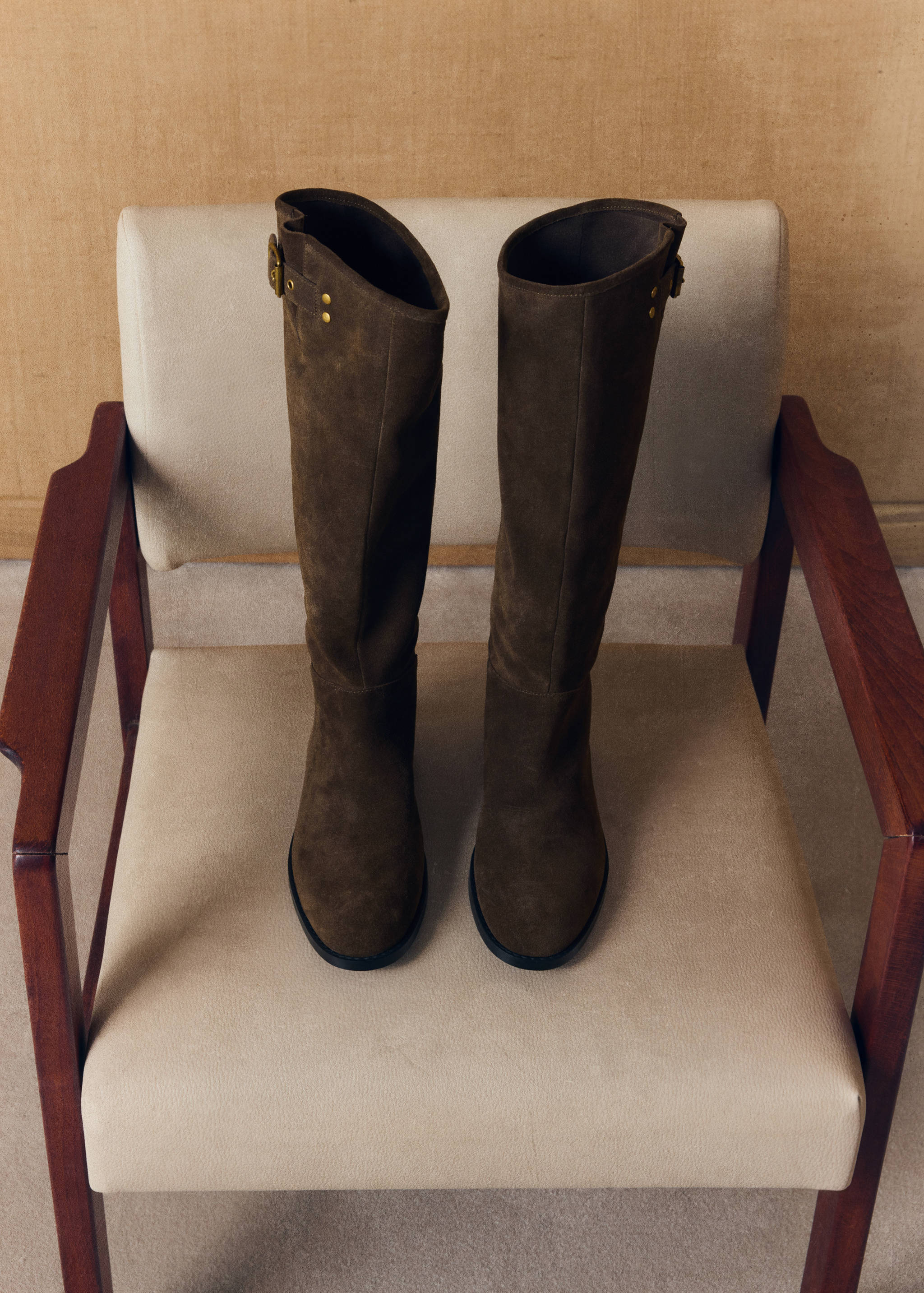 Dark brown suede knee-high boots with buckle and stud details on an armchair.