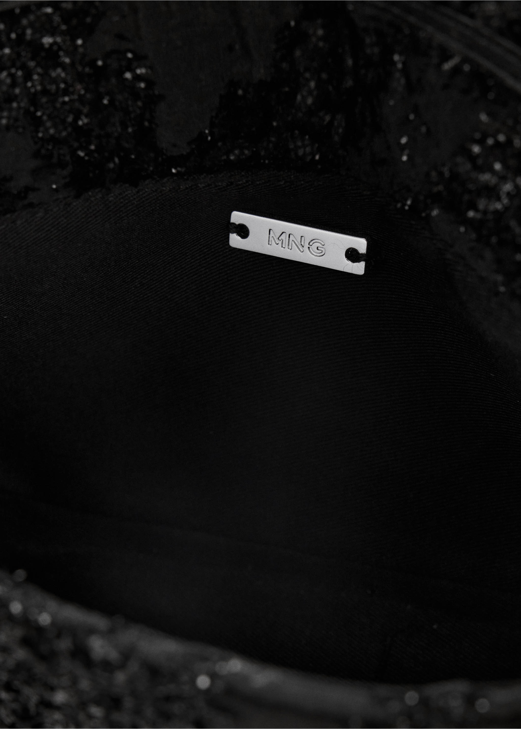 Close-up of a black bandolera bag interior showing the silver MNG metal plaque., Black. Ref: 47051376-00.