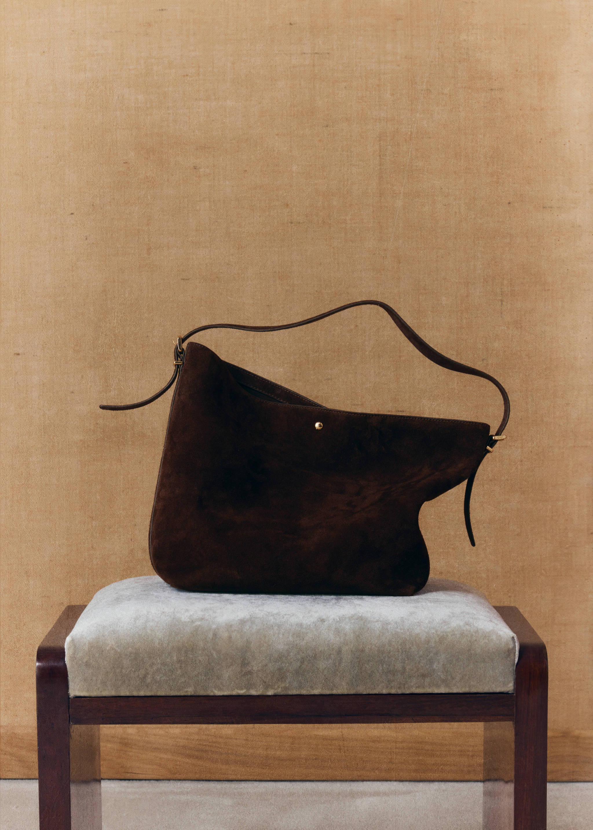 Dark brown suede bucket bag with a slim shoulder strap resting on a stool.