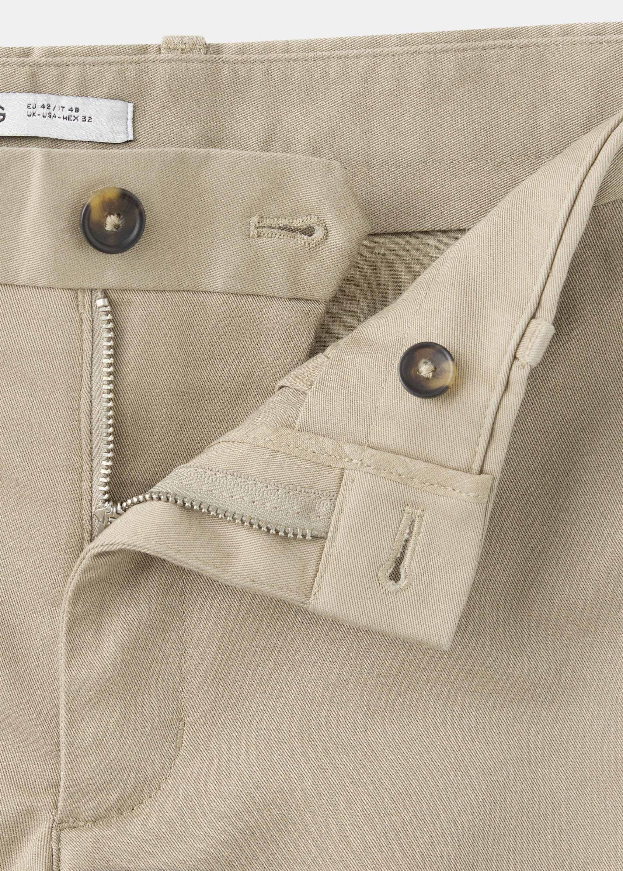 Close-up of beige trousers showing the waistband, zip fly and tortoiseshell button.