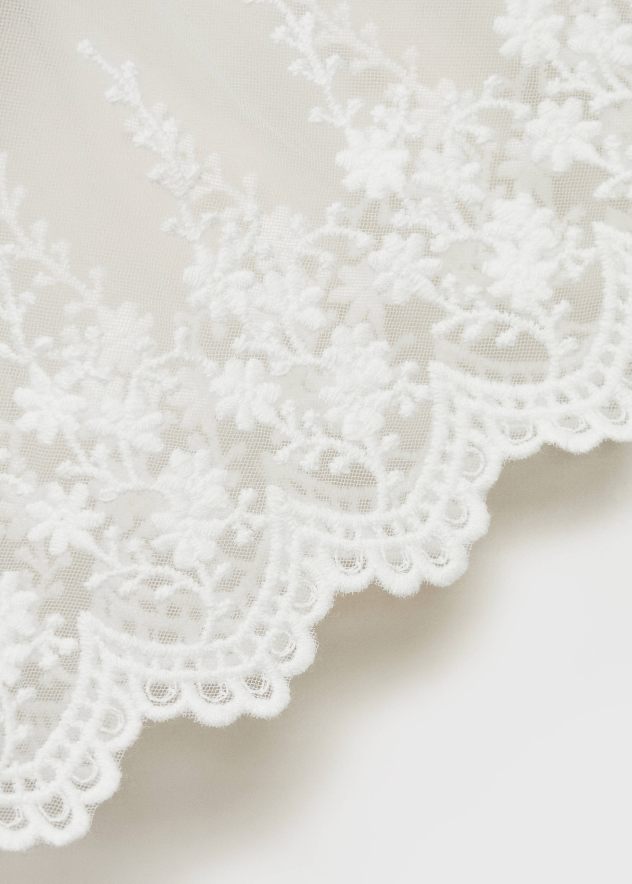 Close-up of a white dress showing sheer tulle with floral embroidery and a scalloped hem.
